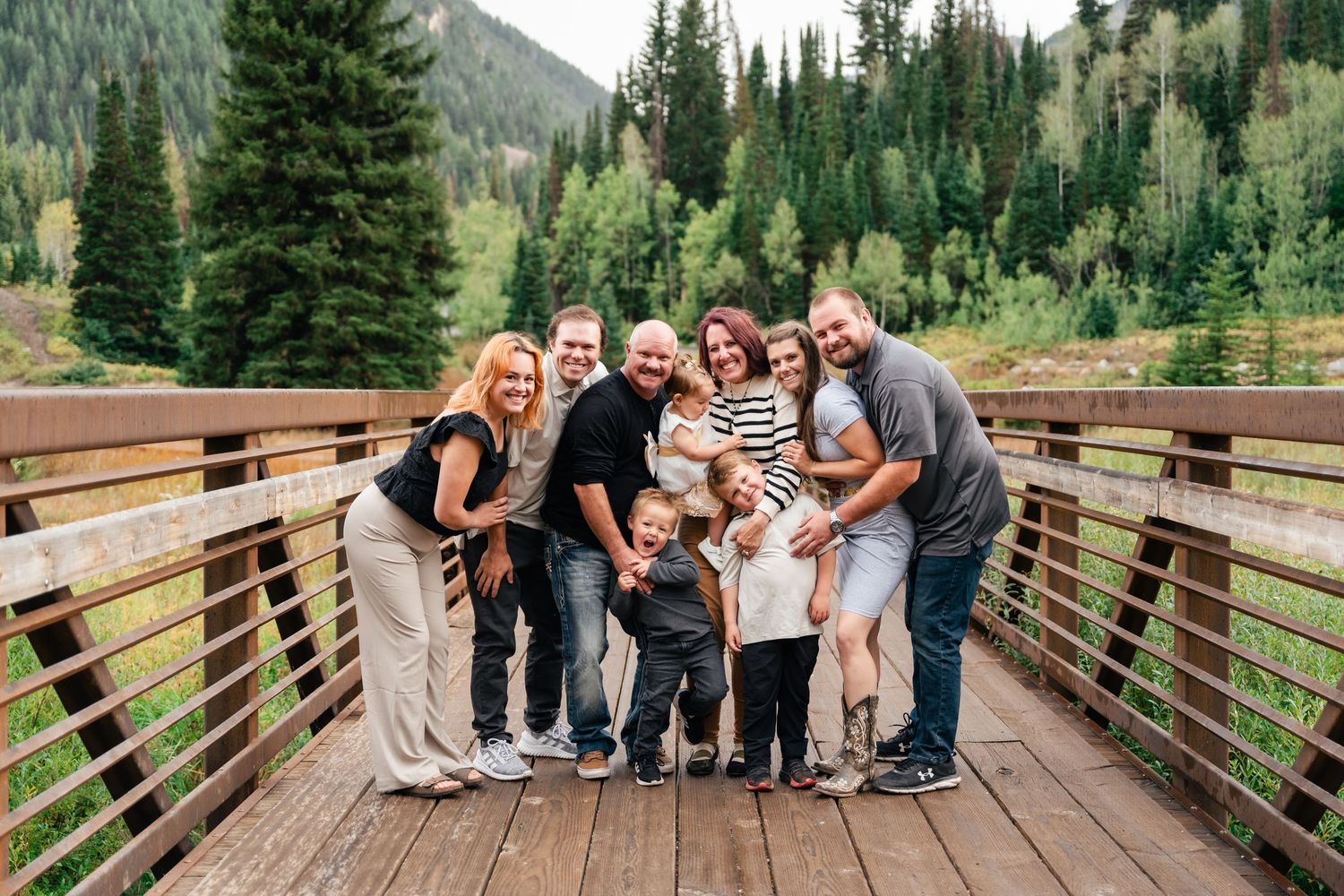 A group of people pose together on a wooden bridge surrounded by evergreen trees in a mountain landscape.