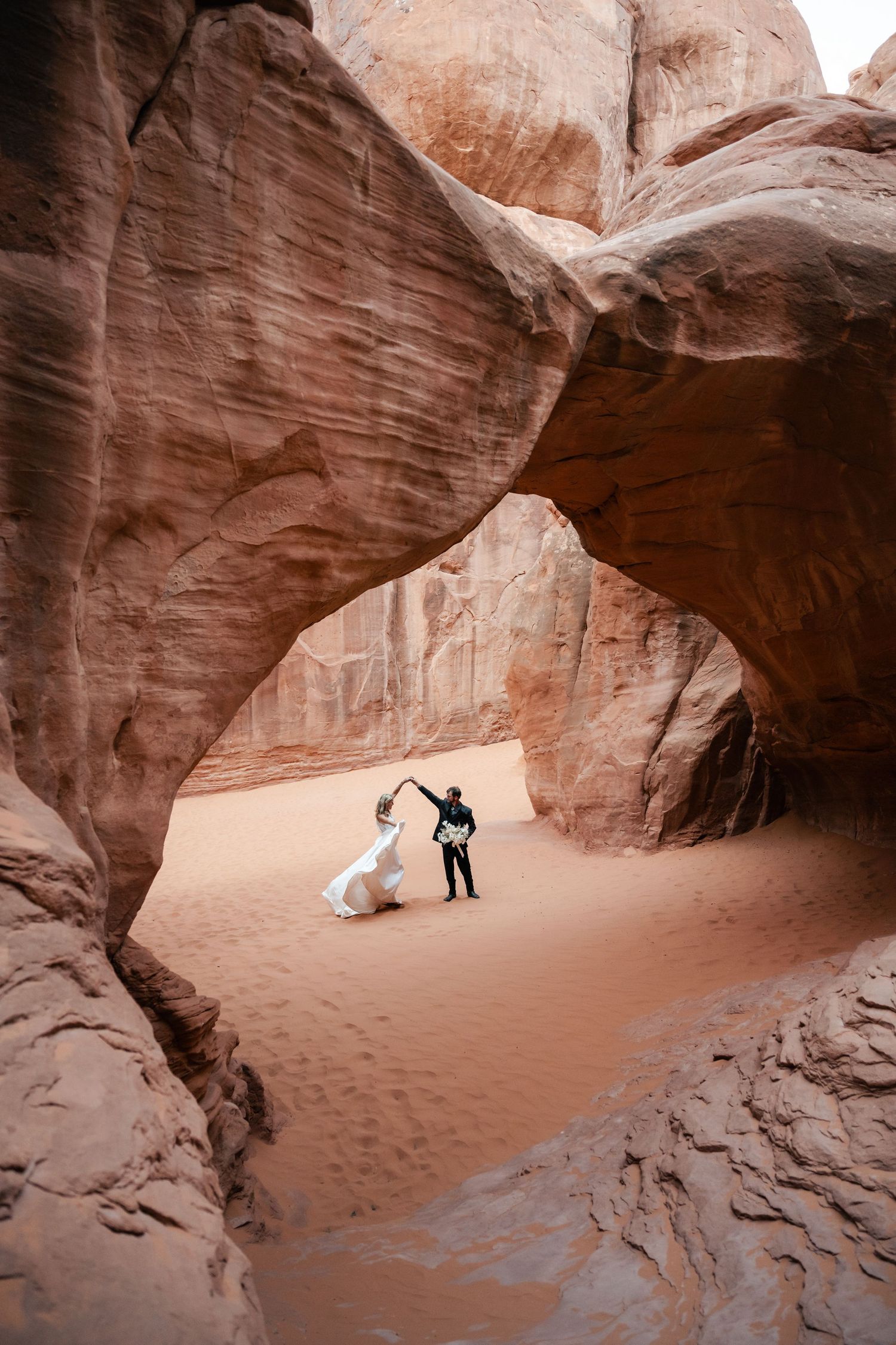 Wedding couple poses beneath towering sandstone arch formations in a desert canyon setting.