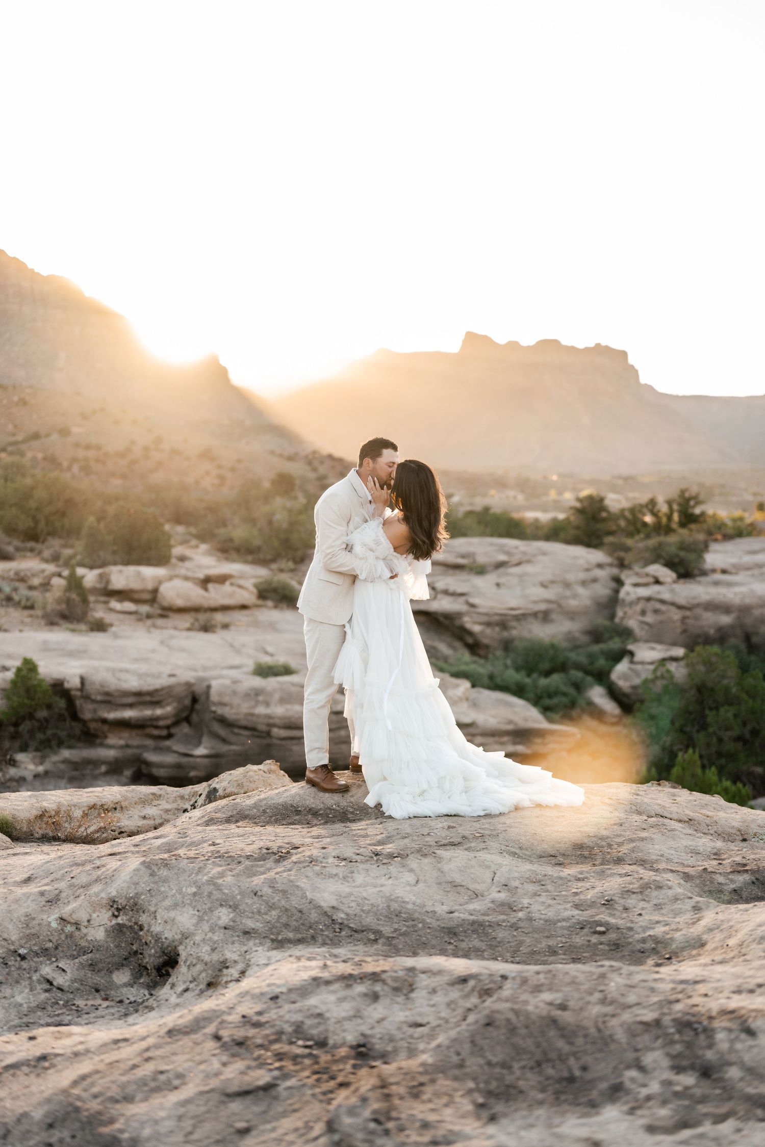 Zion destination photographer. A couple shares a romantic moment on rocky cliffs during golden hour sunset in a desert landscape.