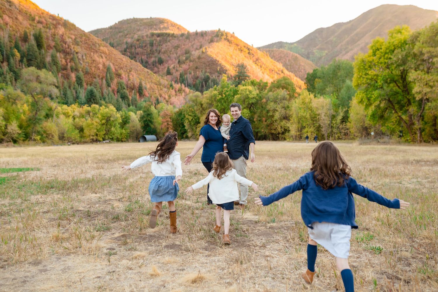 People running joyfully through a golden field with autumn mountains in the background during sunset.