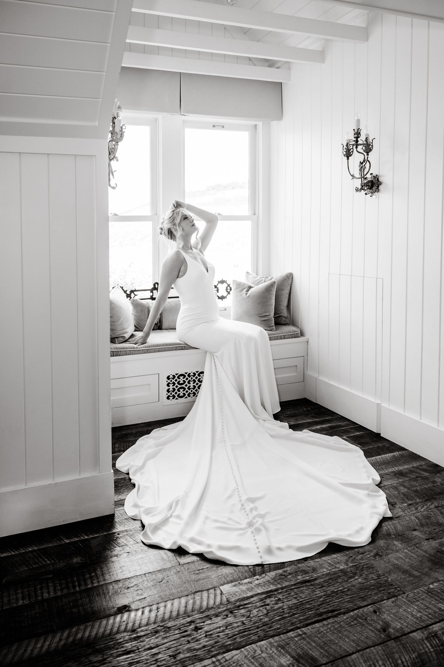 Bride in a flowing white gown sits by a sunlit window seat in a charming white room with rustic hardwood floors in Park City.
