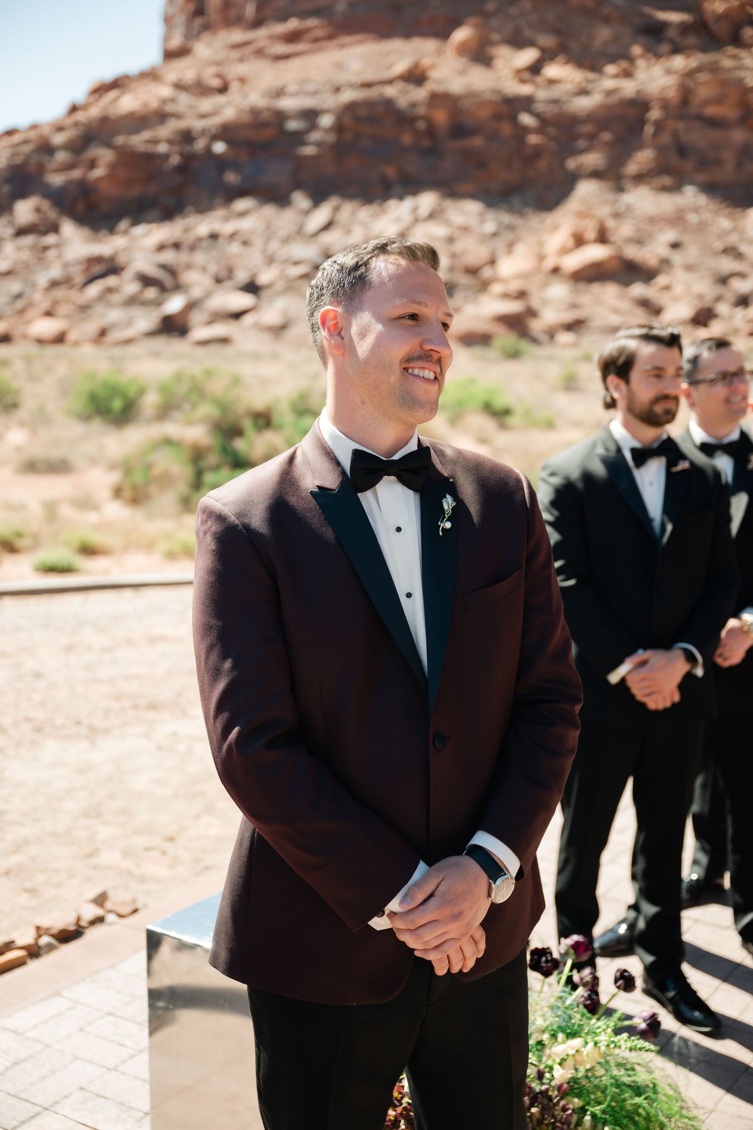 Groom in burgundy tuxedo jacket smiling at outdoor desert wedding ceremony with groomsmen in black tuxedos behind him.
