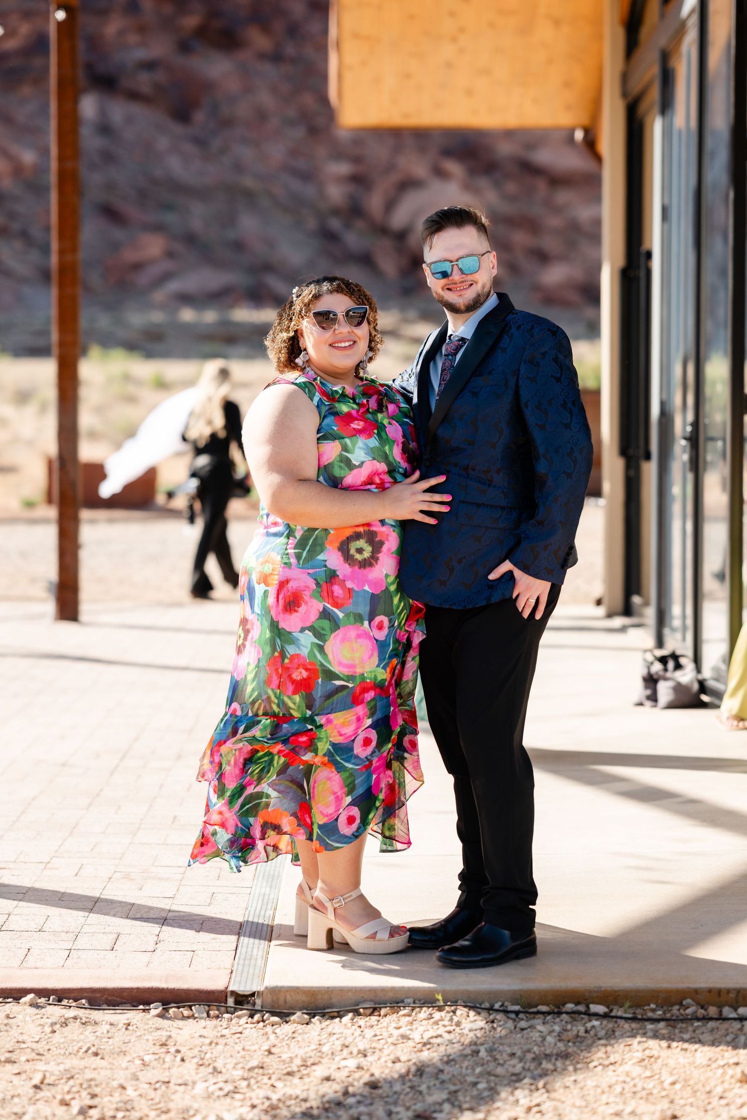 Stylish couple in floral midi dress and navy blazer posing outdoors with red rock canyon background.