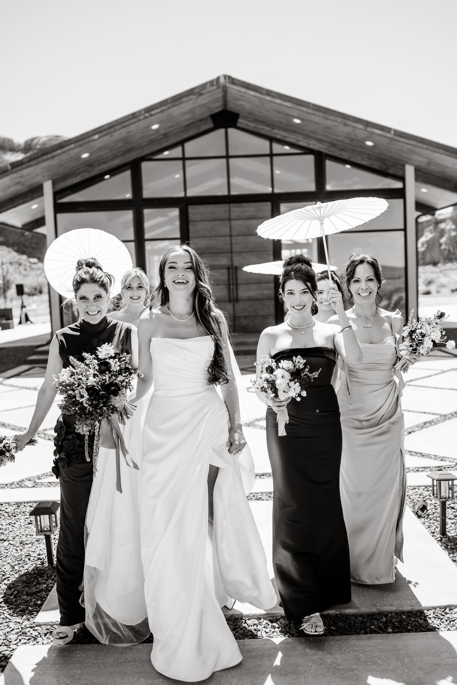 Black and white photo of a bride and four bridesmaids holding parasols and bouquets, walking outside a modern venue.