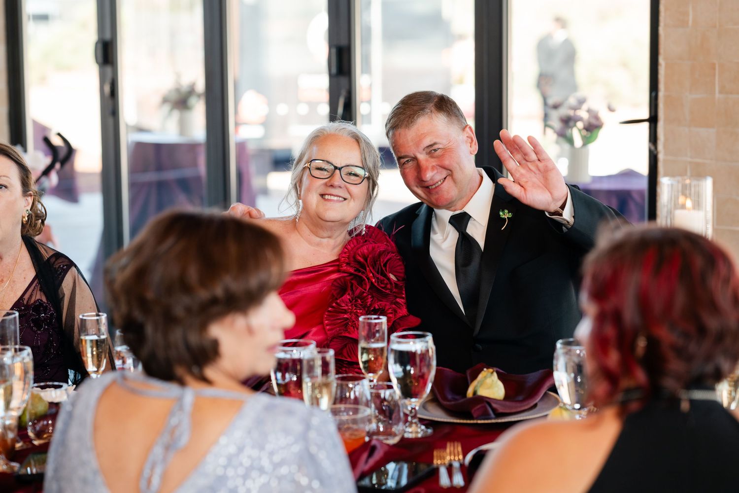Couple smiling and waving at a wedding reception dinner table with champagne glasses and guests in foreground.