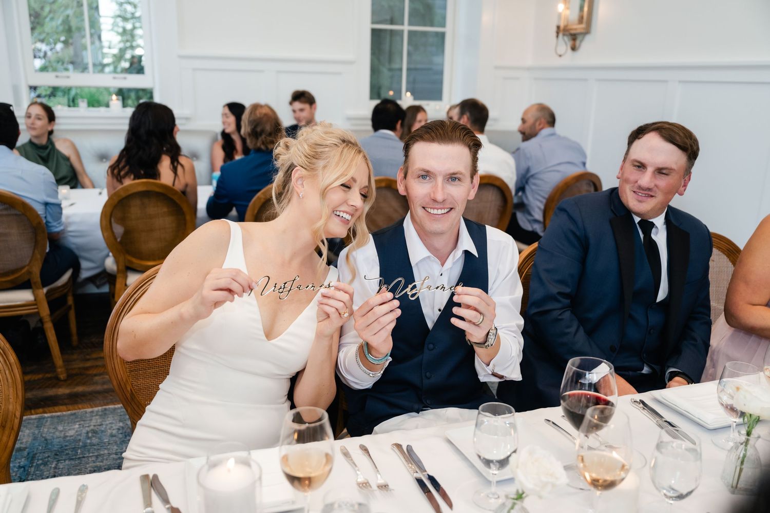 Bride and groom smiling at wedding reception table with guests, holding small items, elegant white dining room.