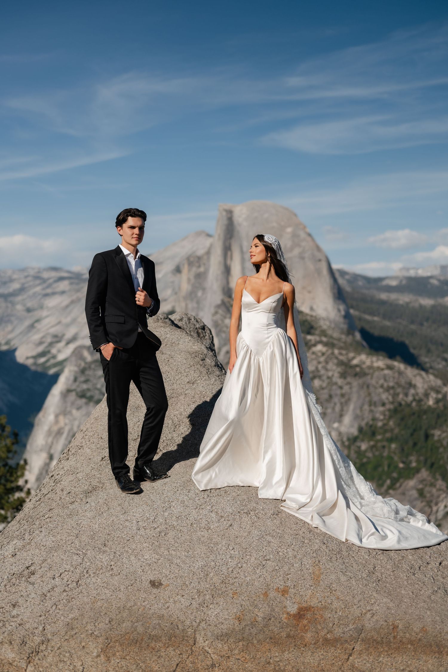 Bride in white gown and groom in black suit stand on Glacier Point with Half Dome in background at Yosemite.
