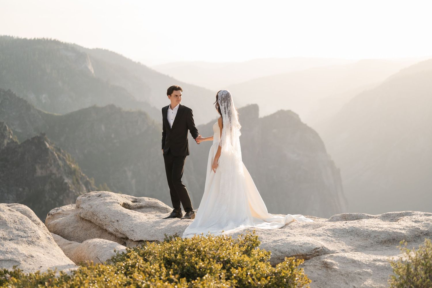 Groom holding bride's hand on sunlit rocky Yosemite overlook with misty mountain valley behind them.