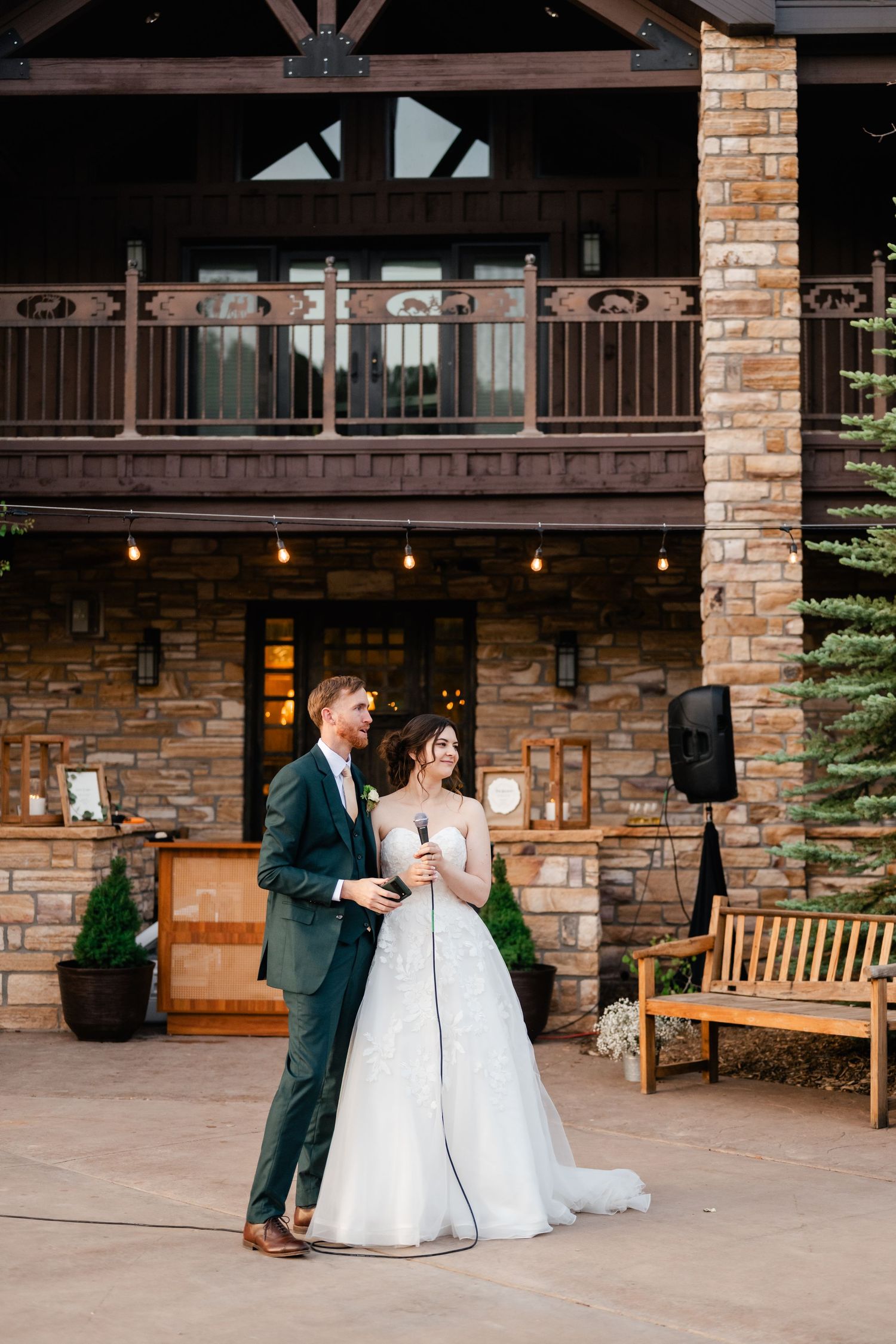 A wedding couple stands together at a rustic venue with brick walls and string lights overhead.