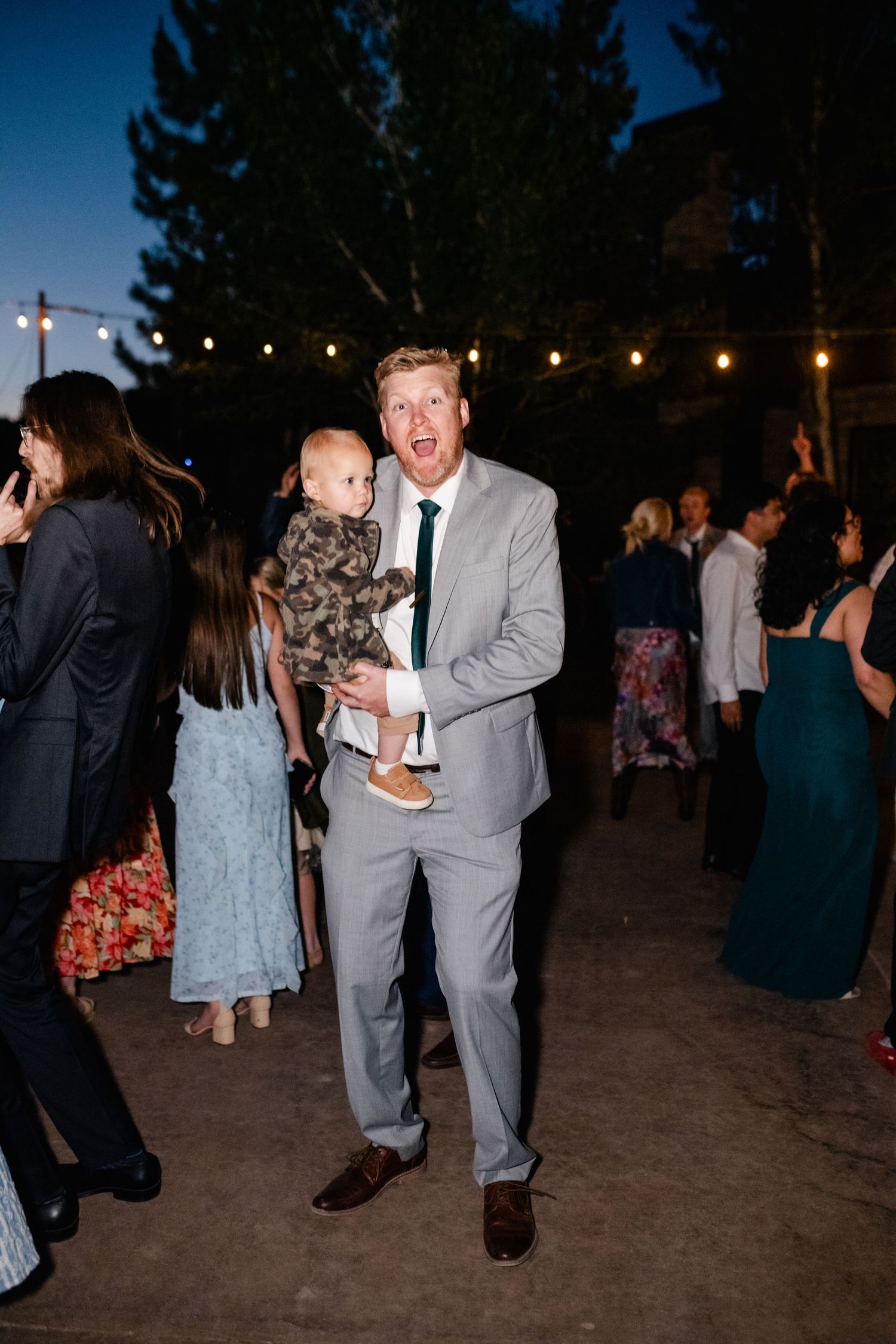 Evening outdoor wedding reception with string lights and guests dancing under dark blue sky.
