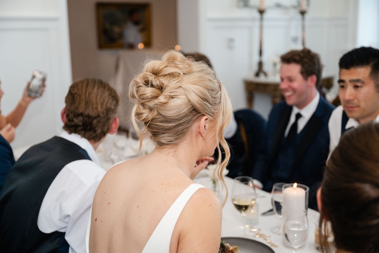 Wedding guests dining and socializing at tables in an elegant room with chandeliers.