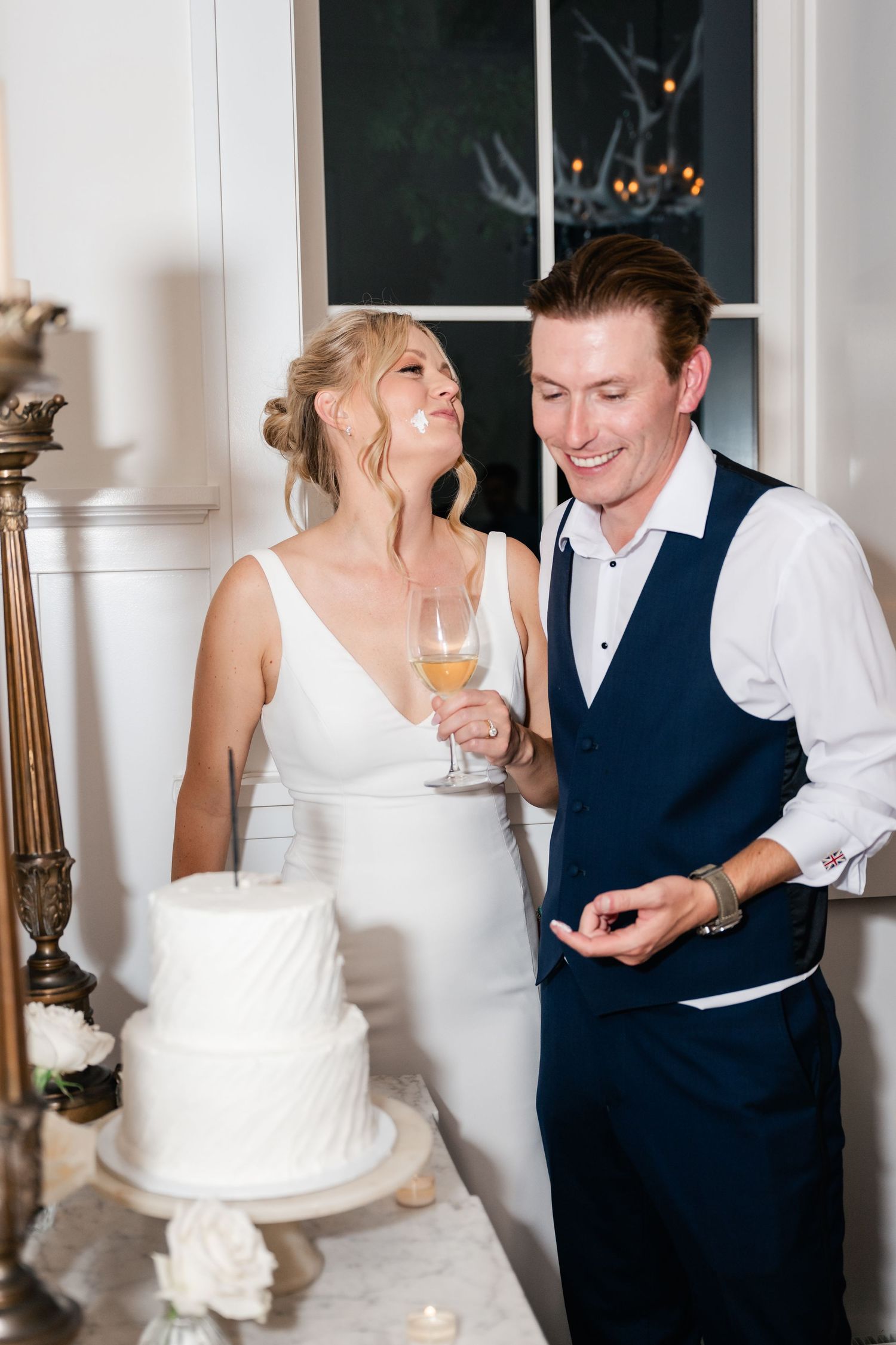 Newlyweds share joyful laughter during their wedding reception beside a white tiered cake.