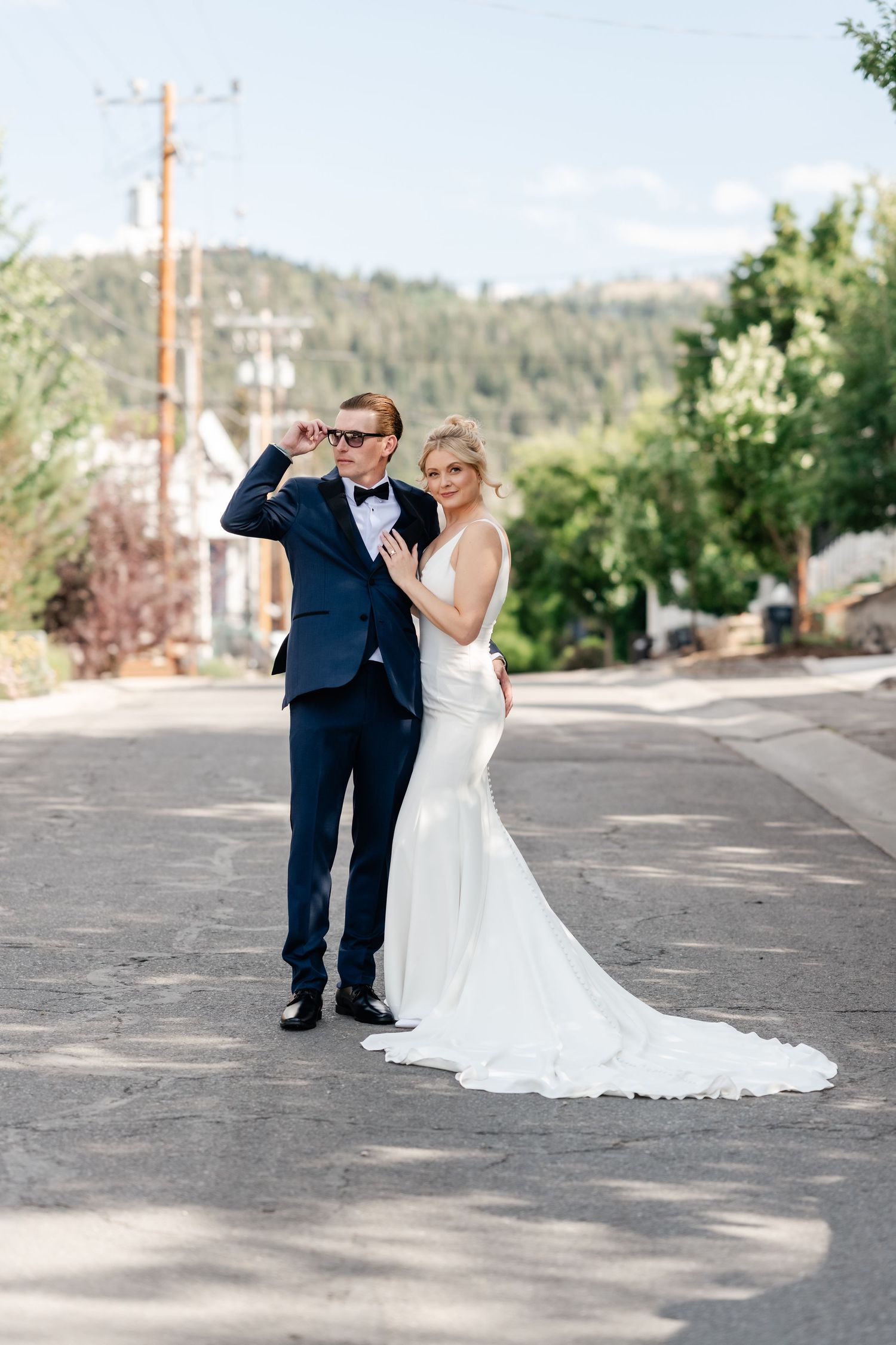A wedding couple poses playfully on a sunny street with mountains visible in the background.