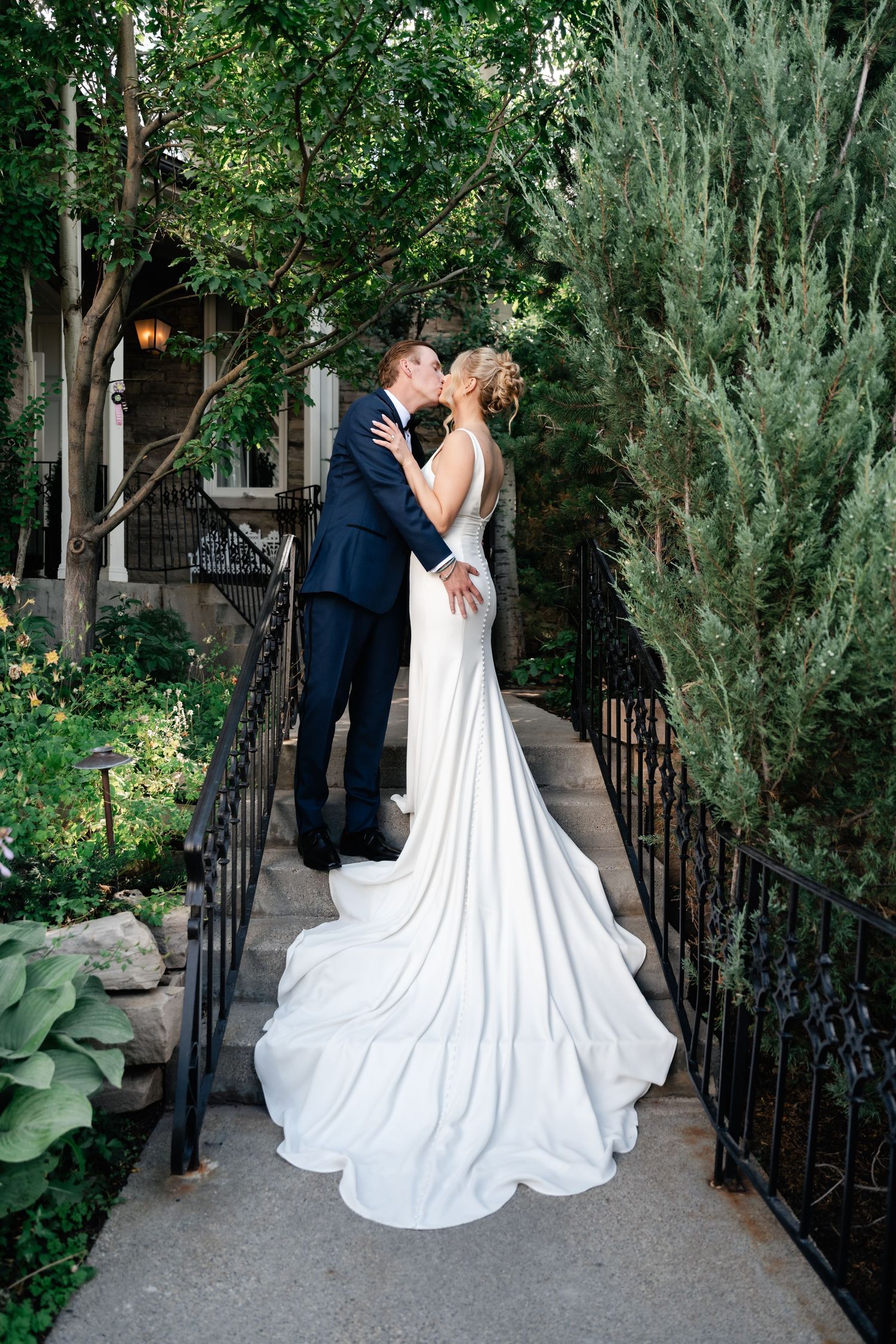 A romantic wedding couple shares an intimate moment in a lush garden setting with wrought iron railings and greenery.