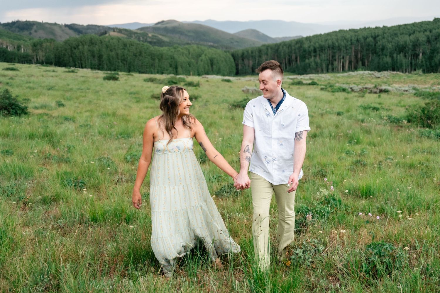 A couple explores a scenic mountain landscape with rolling hills and wildflowers in bloom.
