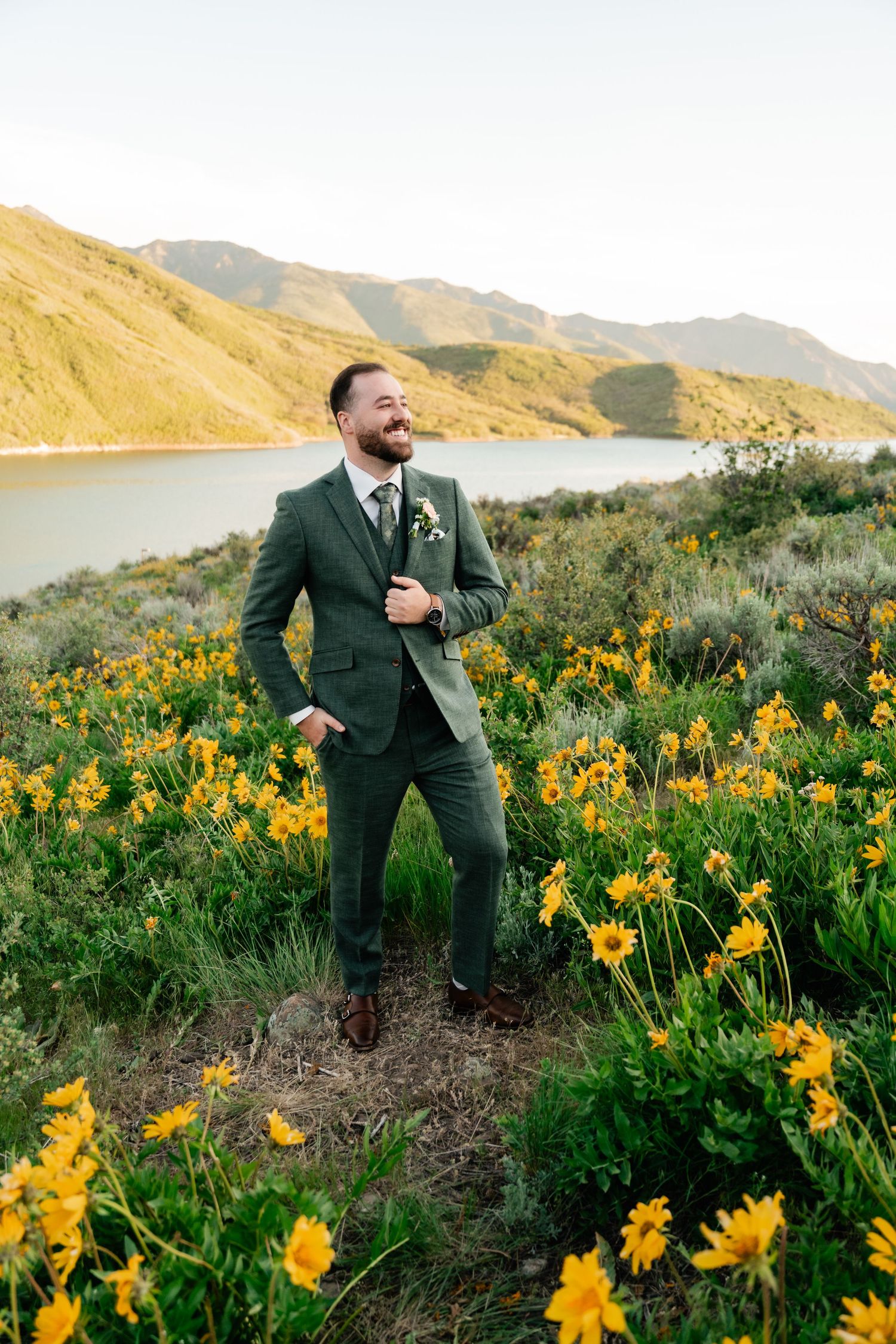 A groom laughs while standing in a field of yellow wildflowers with a mountain lake in the background.