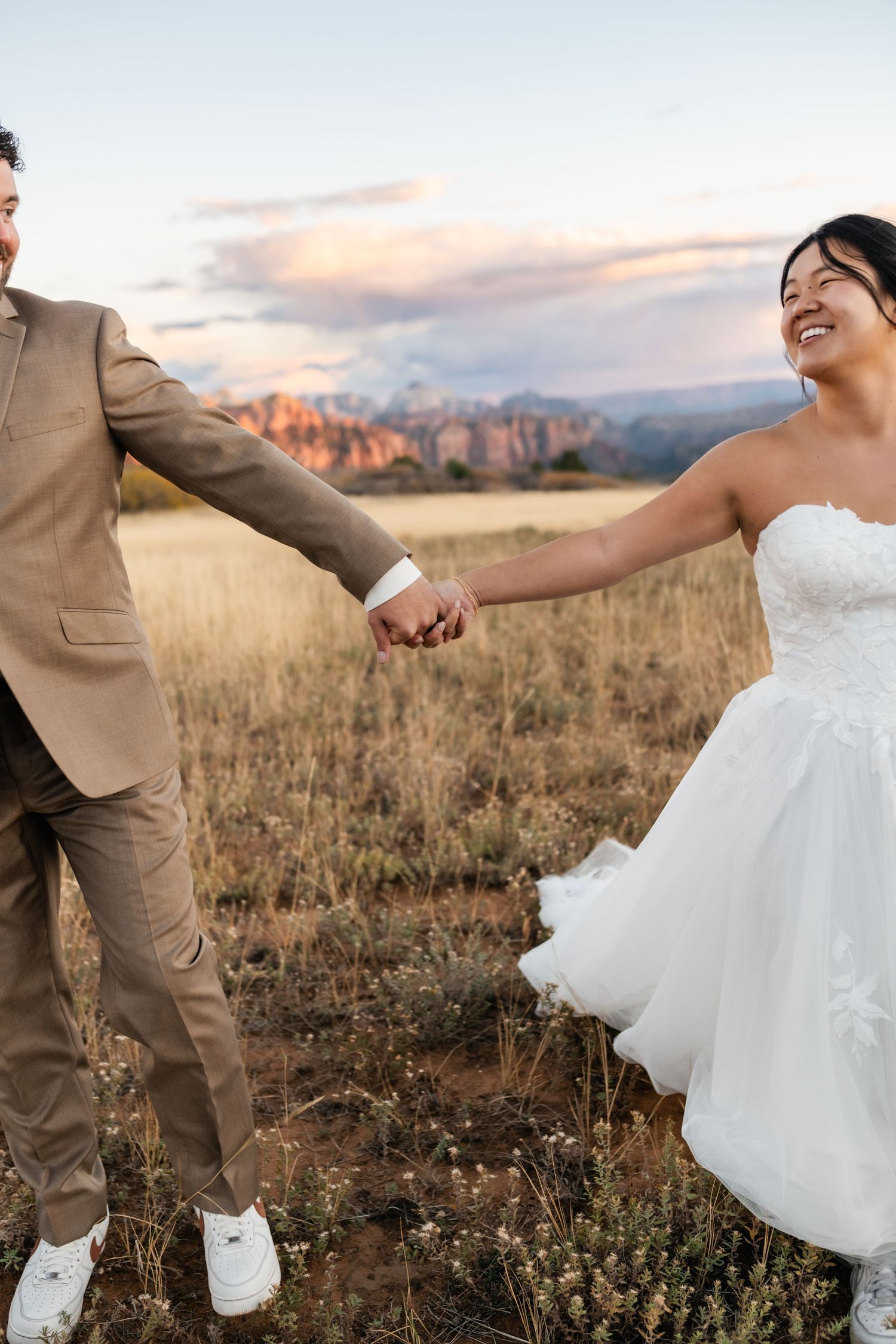 A couple holds hands while walking through tall grass with mountains in the background.
