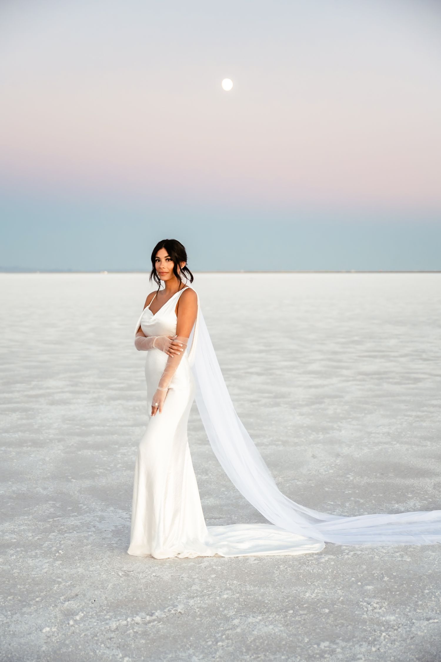 Bride with flowing veil poses gracefully on pristine white salt flats at sunset.