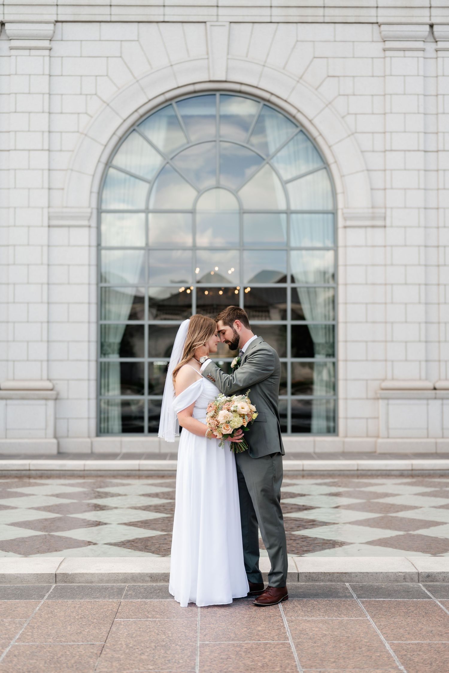 A couple embraces in front of a large arched window on a stone building exterior.