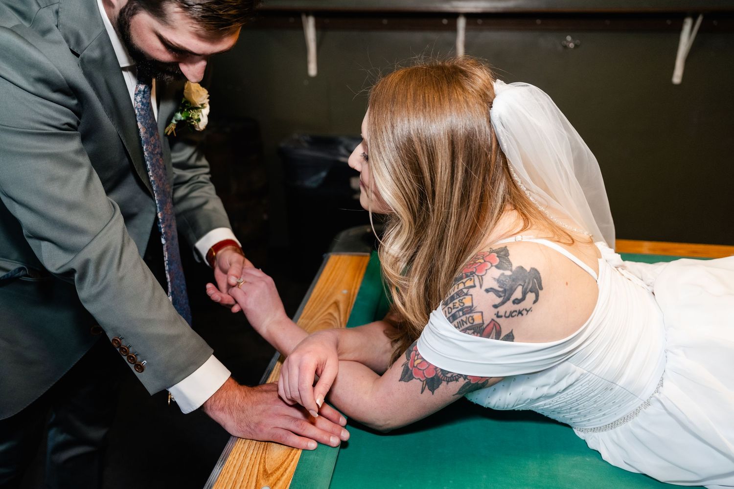 A bride in a white dress shows off her tattoo while leaning on a pool table in a dimly lit bar.