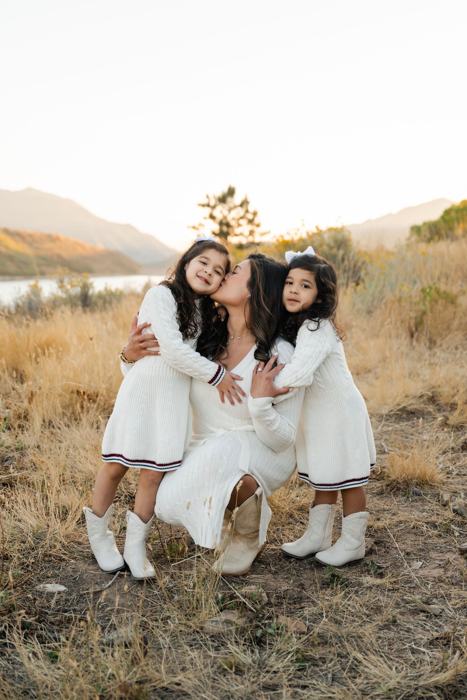 Three people in white outfits share a tender moment on a hillside during golden hour with a lake in the background.