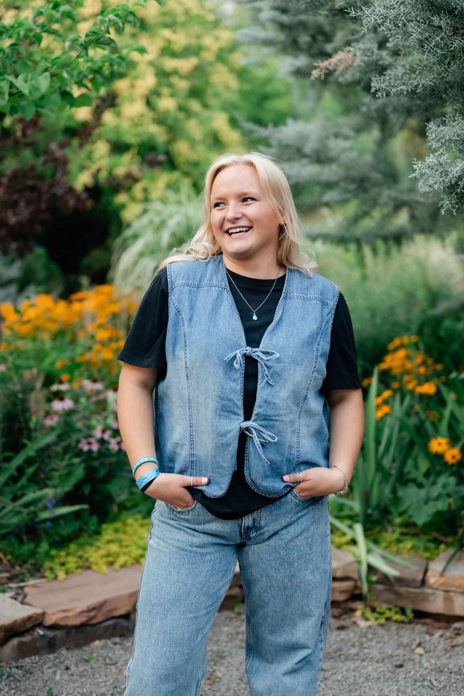 Person in denim vest and jeans stands in front of vibrant garden with orange flowers and greenery.