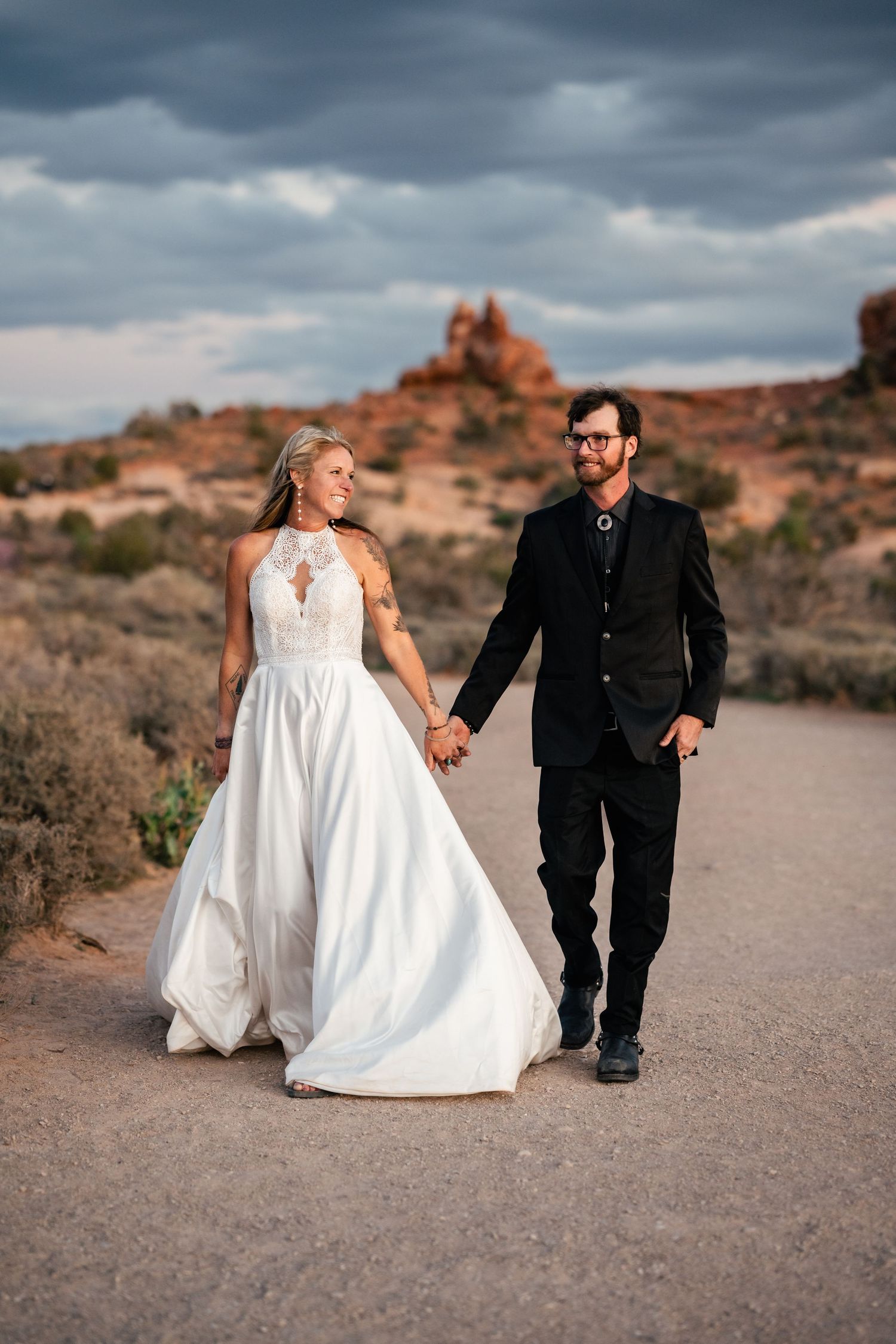 A couple walks together on a desert path with dramatic storm clouds overhead.