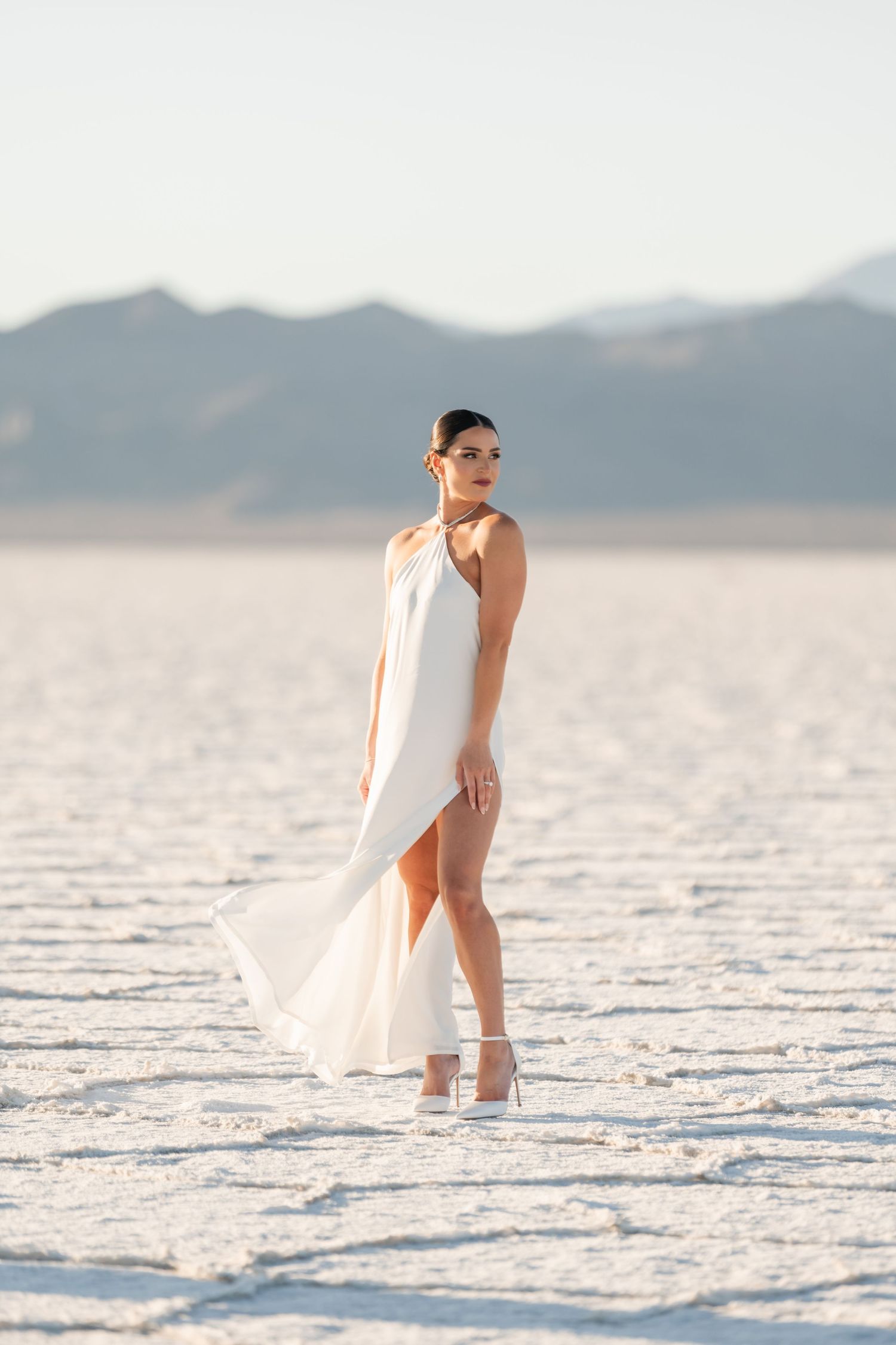 Salt Flats Photos. A figure in a flowing white dress poses gracefully on a stark white salt flat landscape with mountains in the distance.