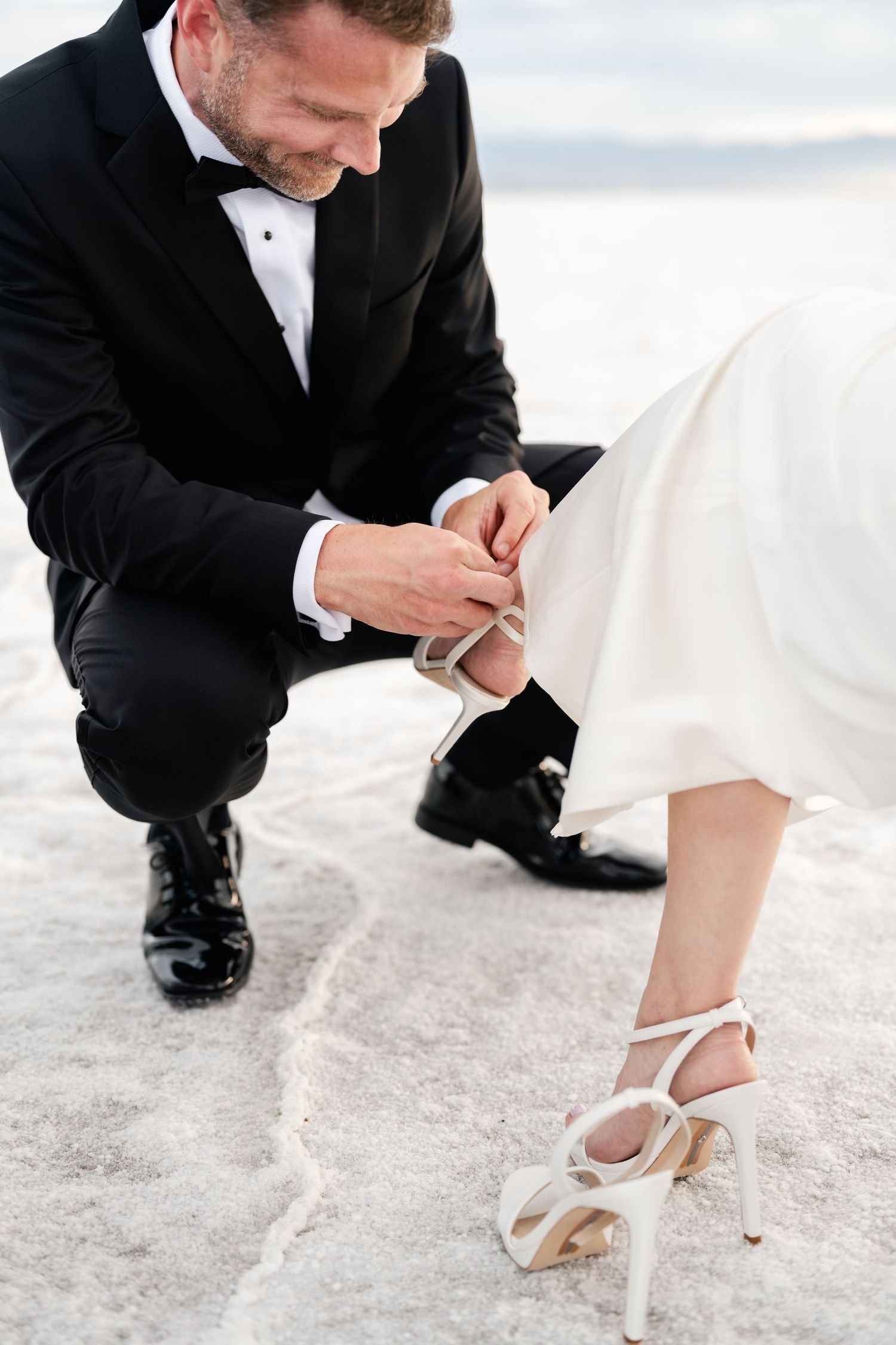 Salt Flats Elopement Photographer. A groom in a black tuxedo kneels down to help adjust a bride's elegant white high-heeled shoe.