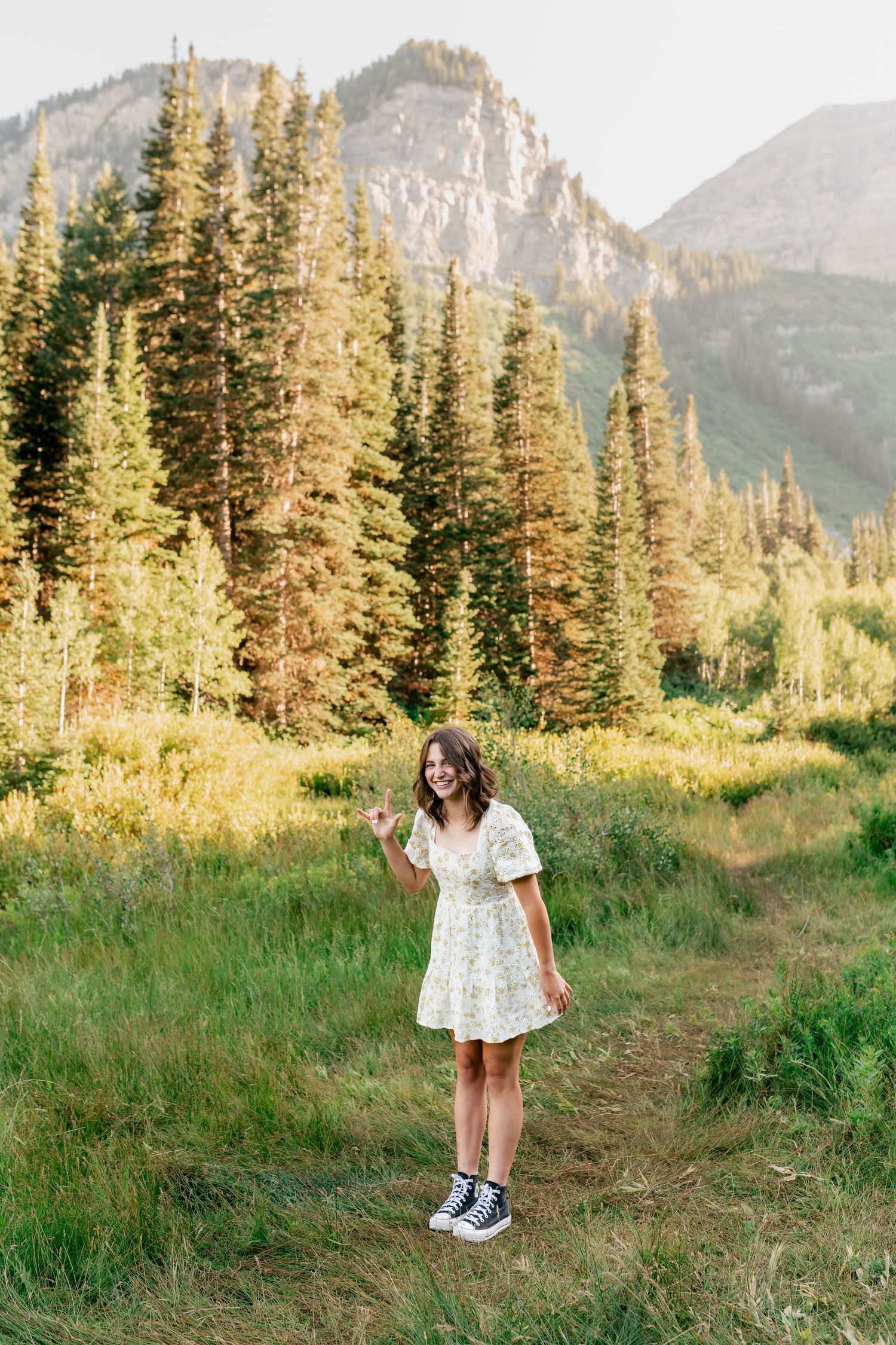 A figure in a white dress poses against a backdrop of majestic mountain peaks and evergreen trees.