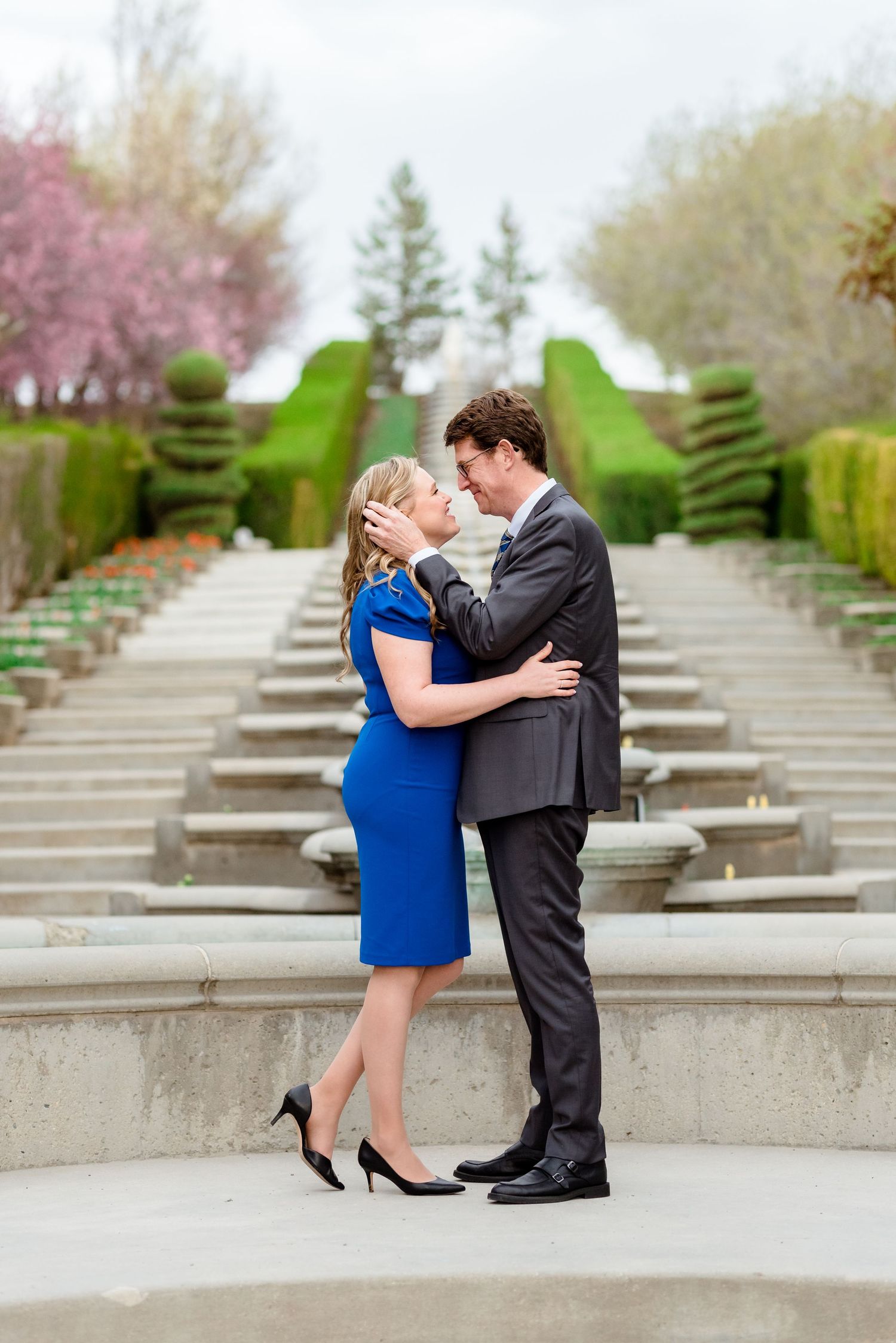 A formal couple embracing on garden steps with manicured hedges in the background.