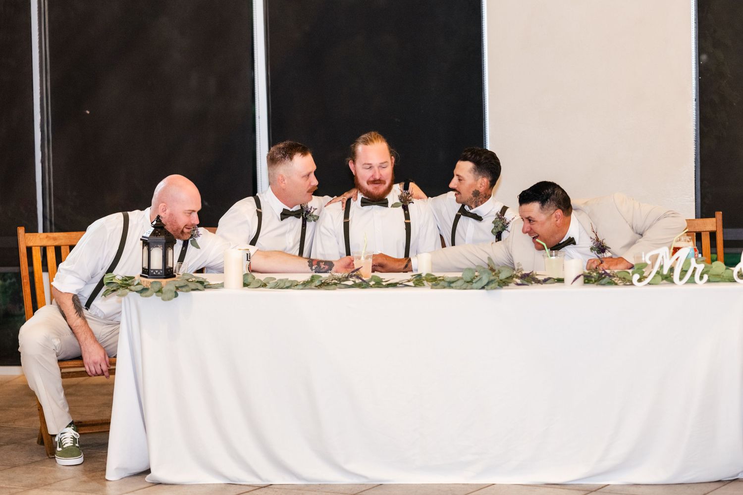 Groomsmen in white shirts and suspenders lean in laughing together at a wedding reception head table with greenery garland.