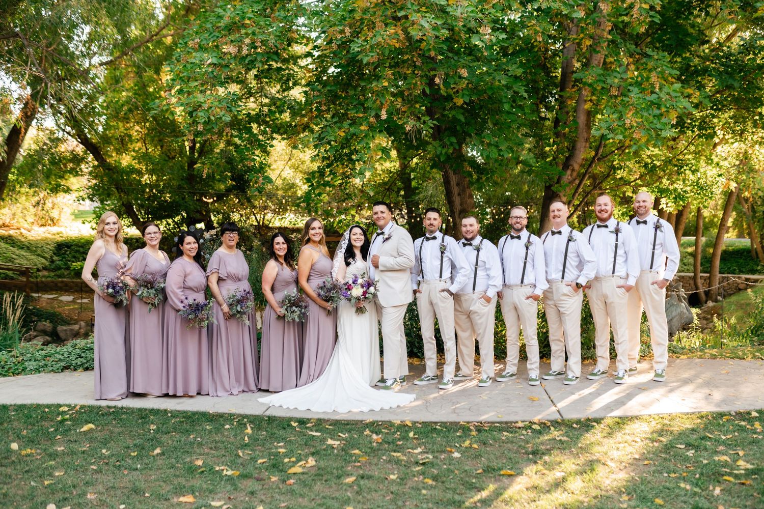 Wedding party in mauve dresses and khaki suits poses together on garden path in sunlit outdoor setting.