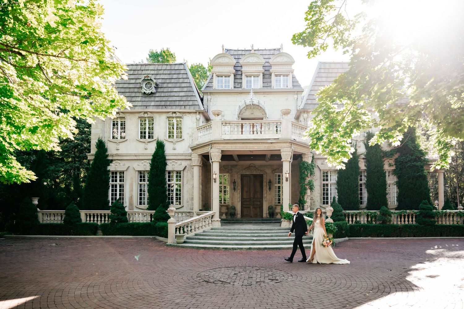 Sunlit wedding portraits in front of a grand white mansion with manicured gardens.