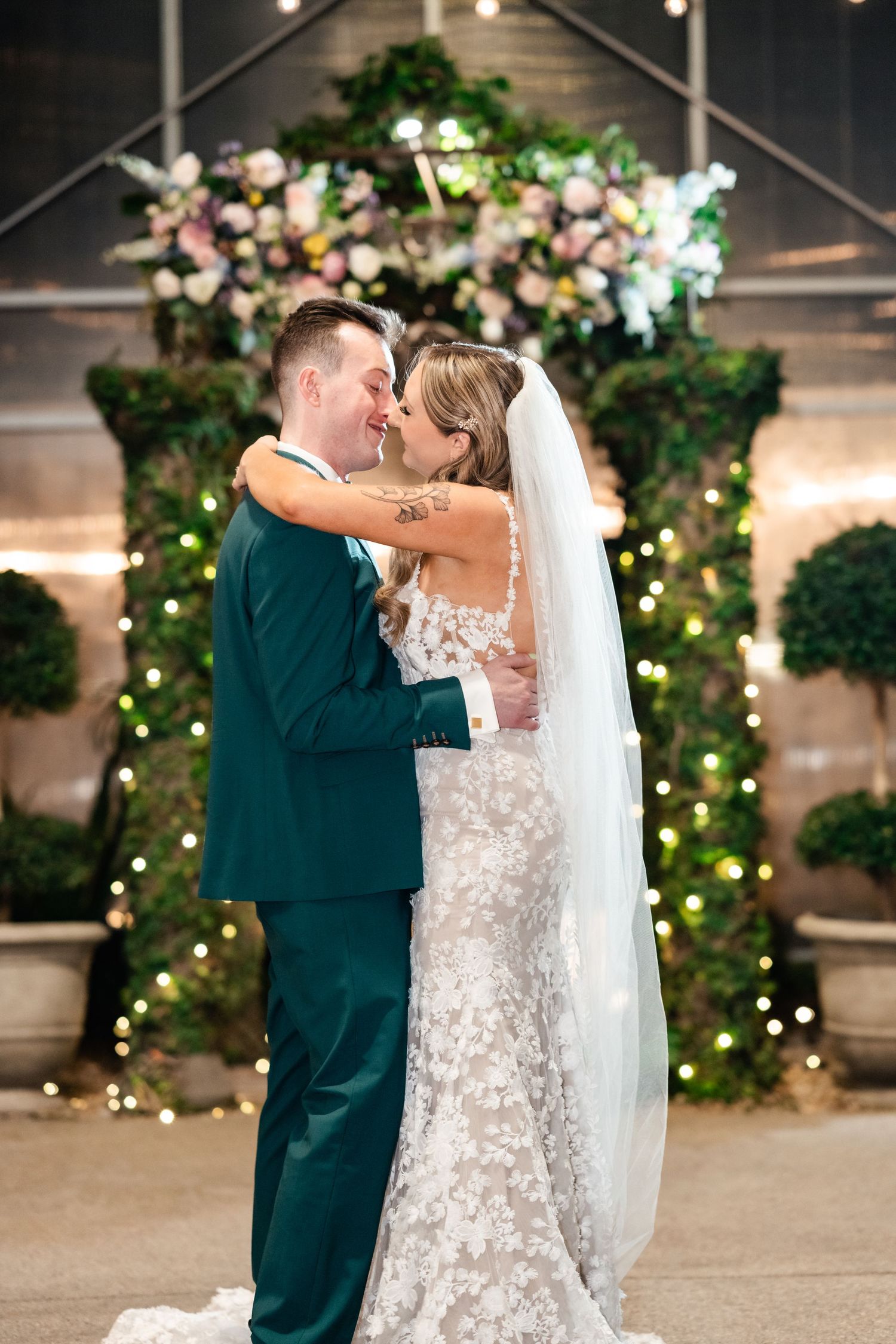 A couple shares their first dance at their wedding reception under a floral arch decorated with twinkling lights.