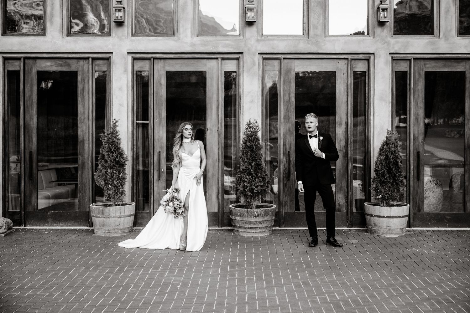 A black and white wedding photo showing a couple standing apart in front of industrial brick doorways with potted plants.