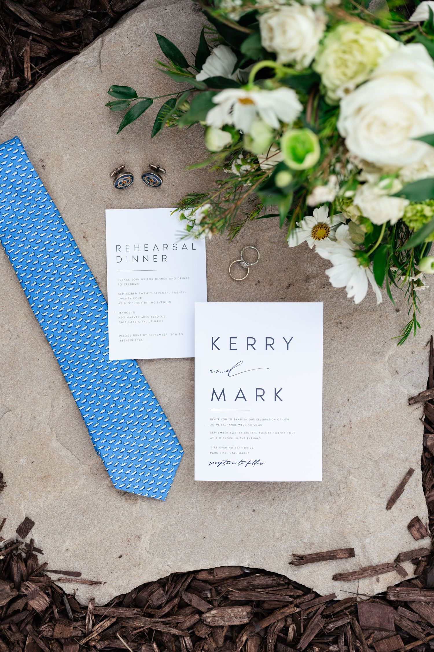 Wedding details including white floral arrangements, blue tie, name cards and rings on a gray surface.