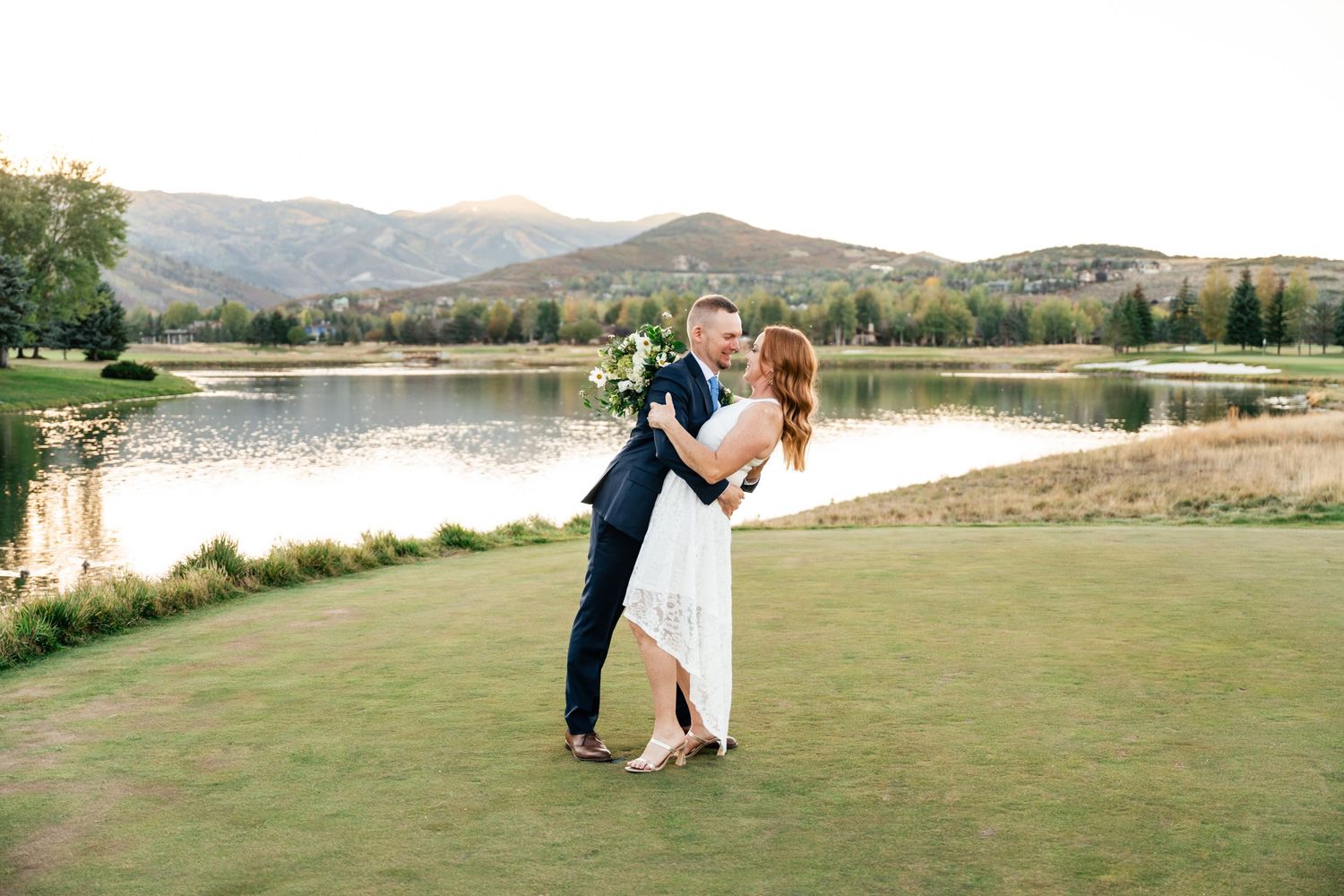 Couple embraces during romantic sunset photoshoot on grassy lawn overlooking scenic mountain lake.