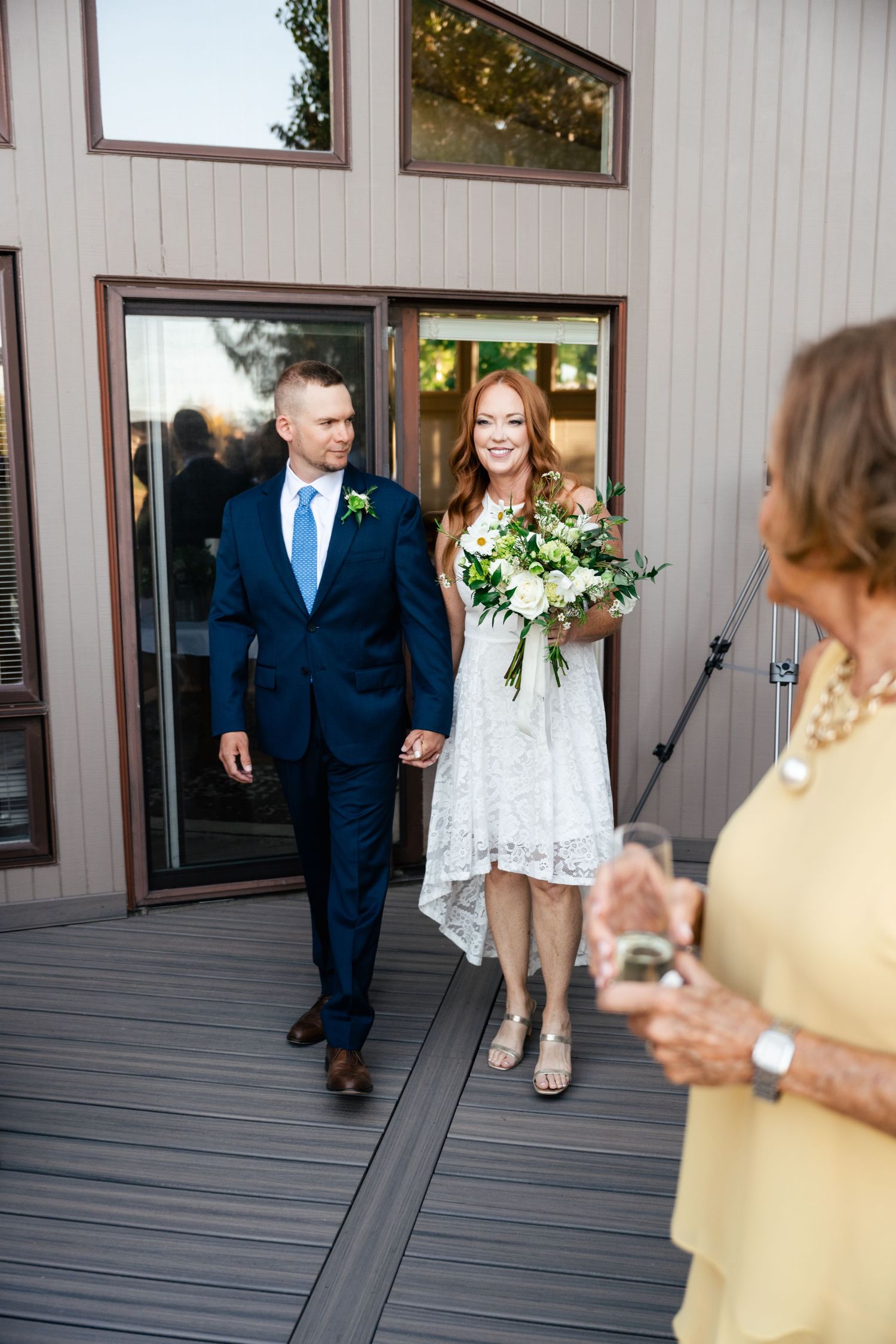 A couple exits through glass doors onto a gray deck during an outdoor wedding ceremony.