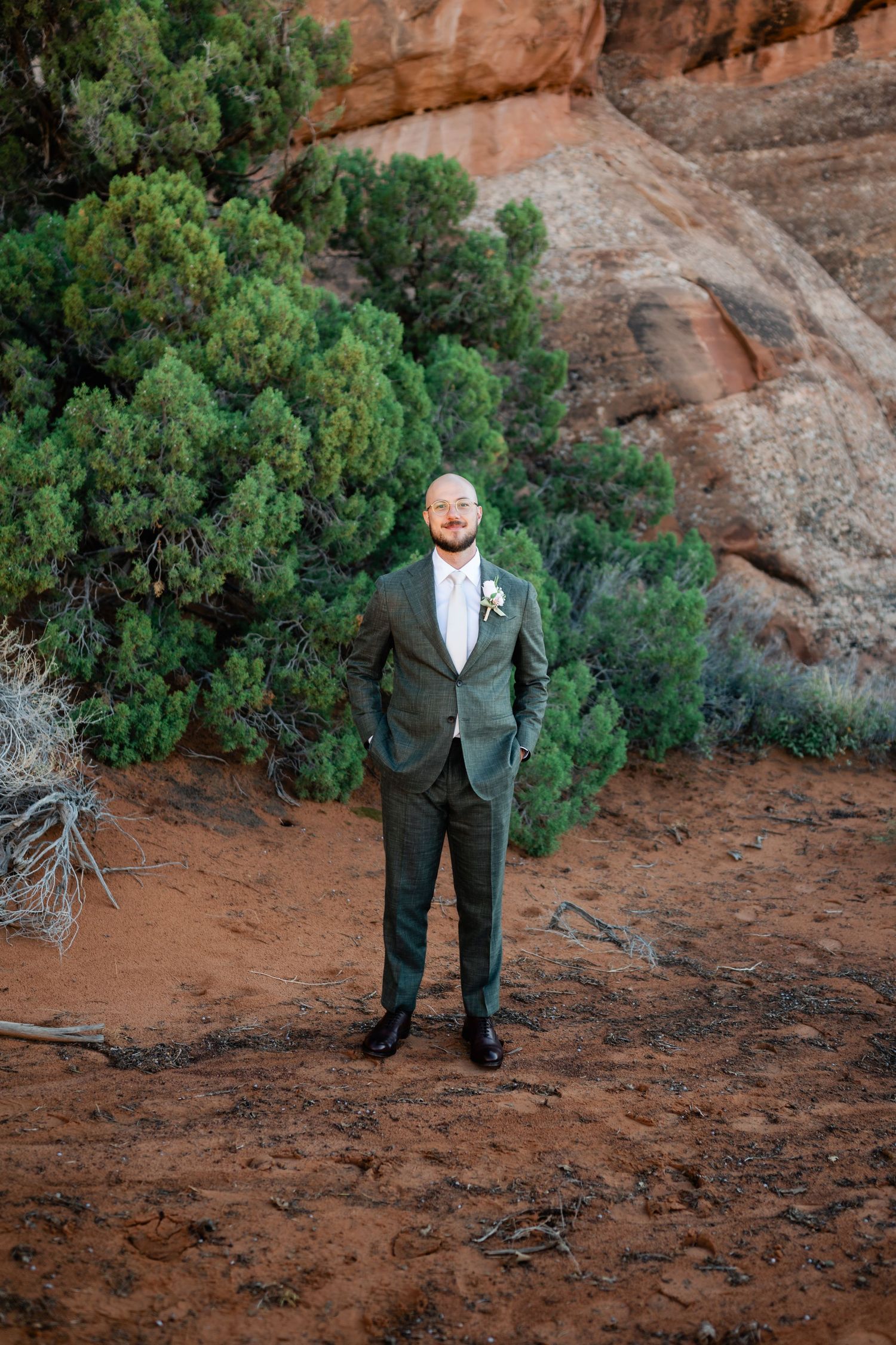 A couple takes wedding photos against stunning red rock formations and desert brush in a scenic outdoor setting.