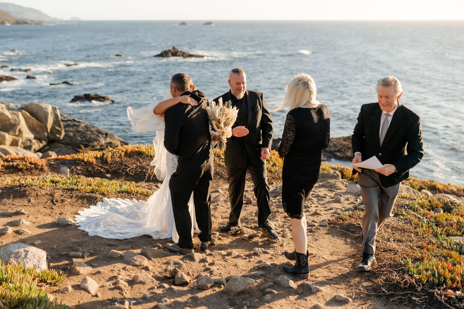Small wedding group gathers on rugged coastal cliffs during intimate outdoor ceremony.