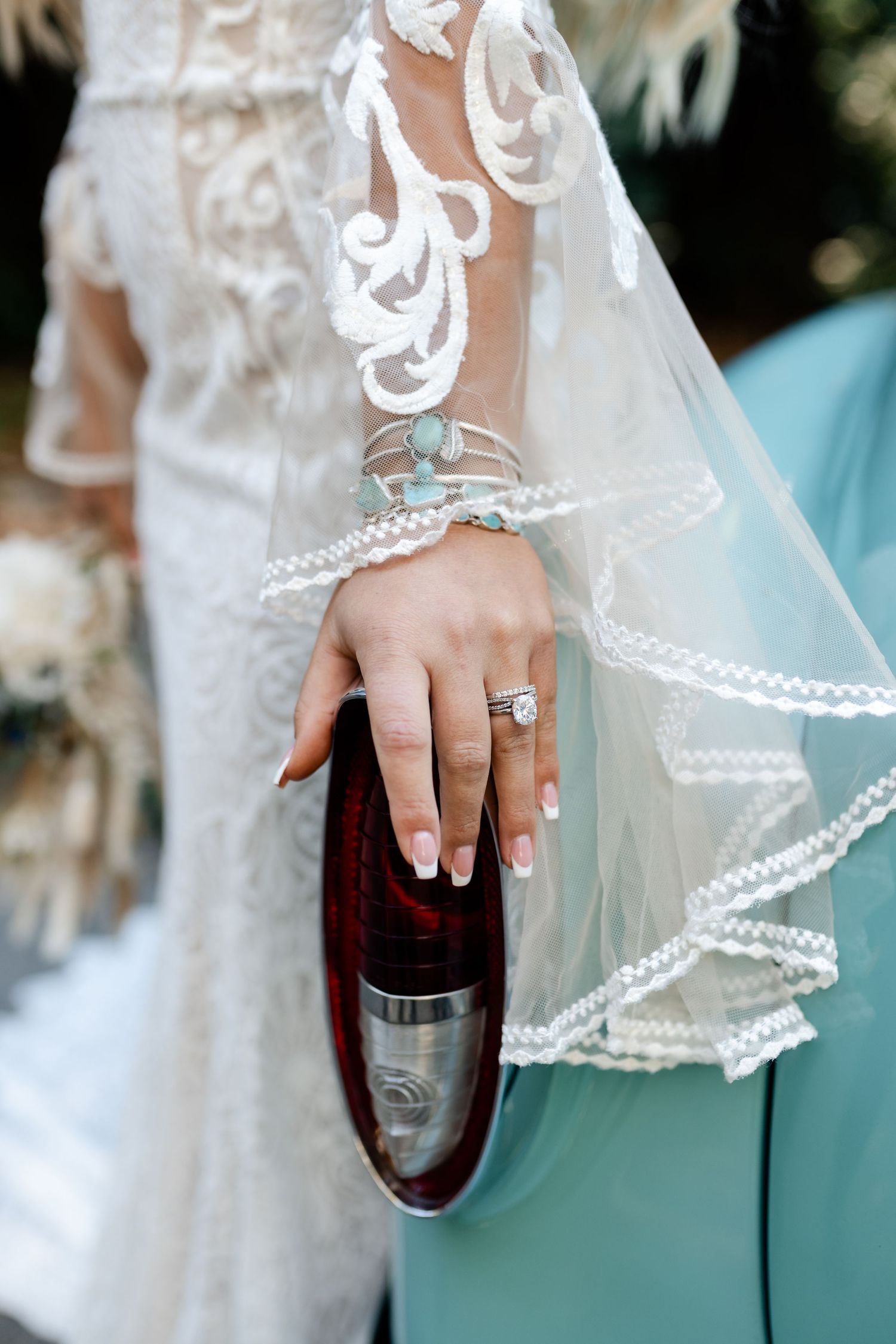 Close-up detail of delicate lace wedding dress sleeve and engagement ring holding a wine bottle.