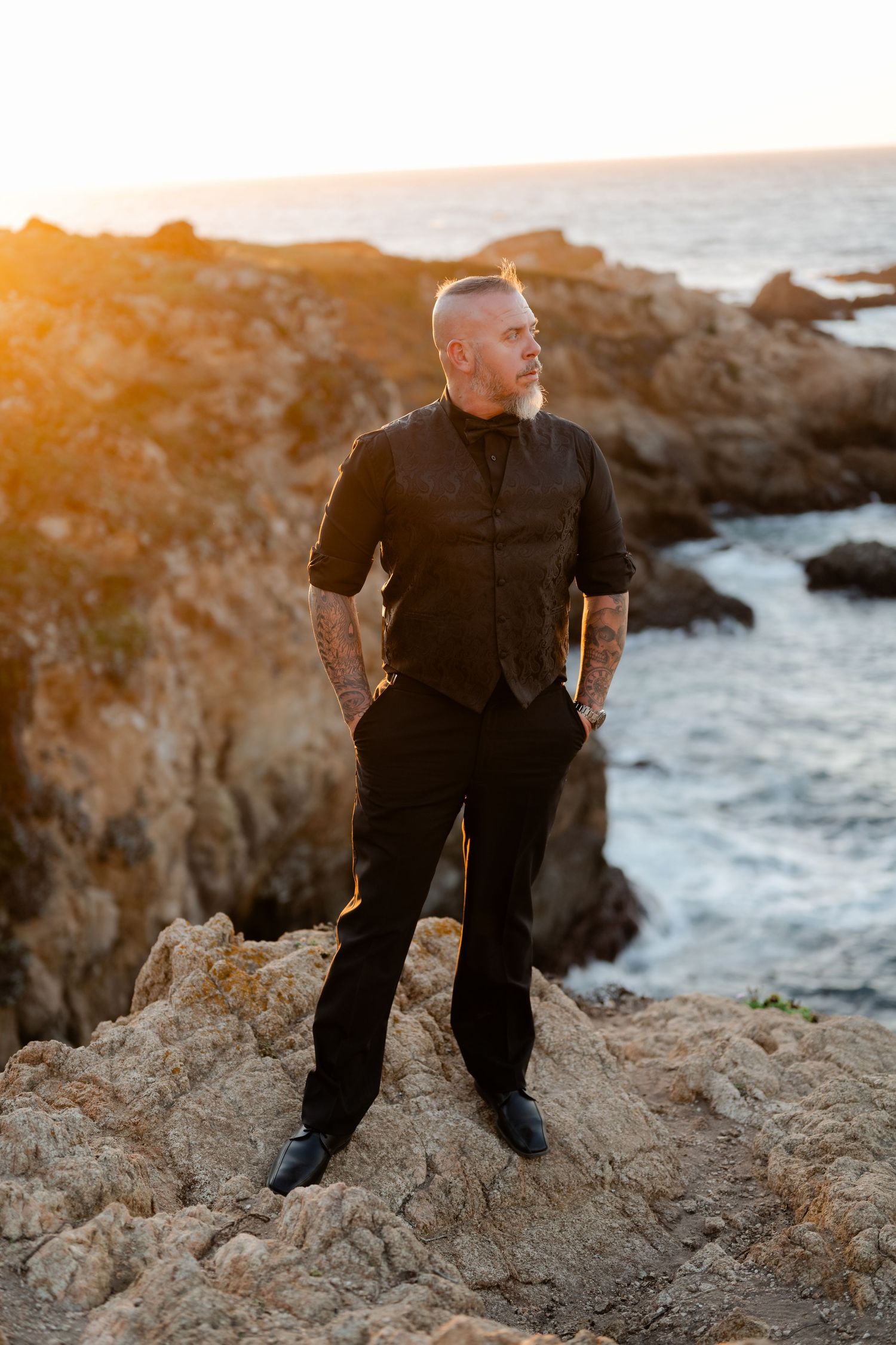 Person in black clothing sits on rocky coastal cliffs at sunset with ocean waves crashing below.