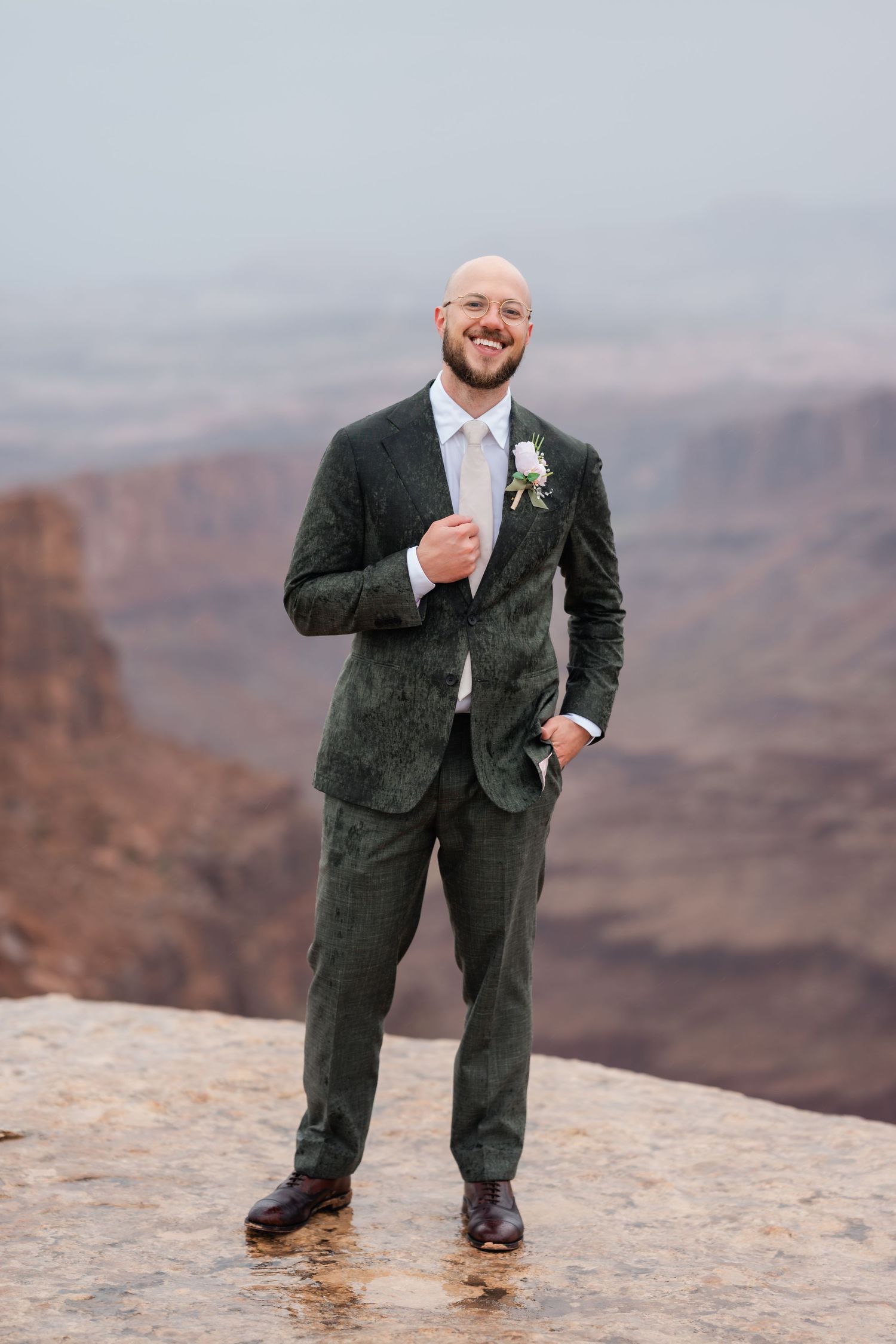 A person in a dark suit poses on the edge of a scenic canyon overlook with dramatic mountain views in the background.
