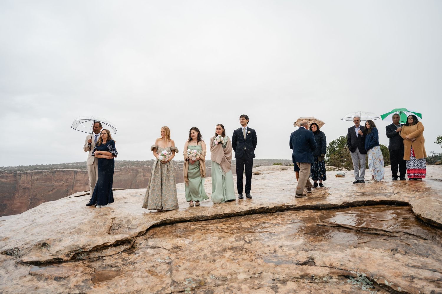 Wedding party gathered on rocky cliff edge overlooking canyon landscape.
