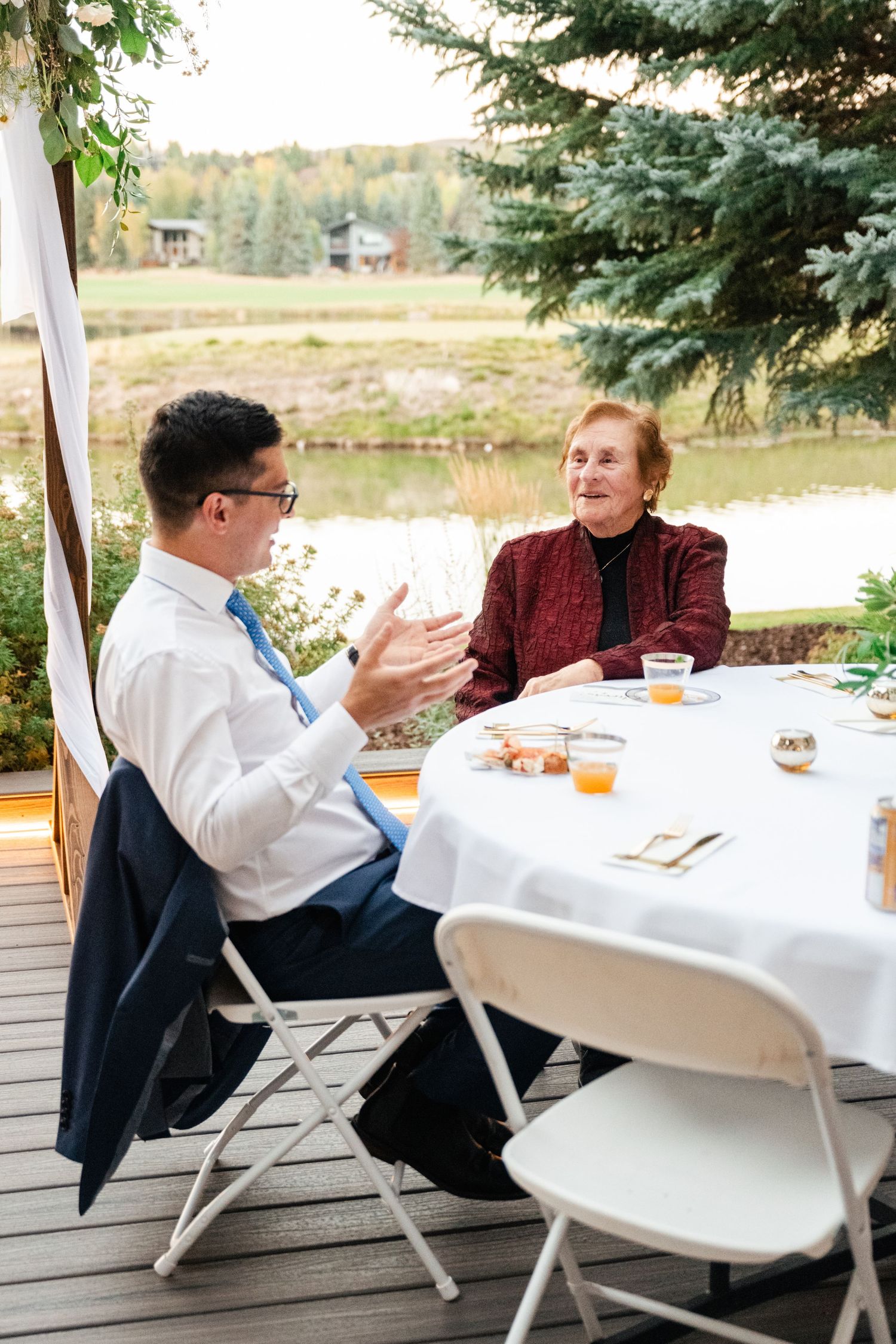 Guests dining at white-clothed tables on an outdoor deck overlooking a scenic lake.
