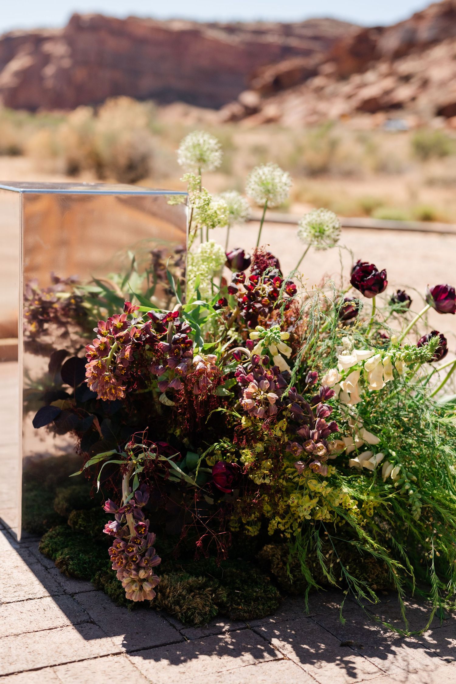 Dark floral arrangement with burgundy blooms and allium against red rock desert landscape backdrop.