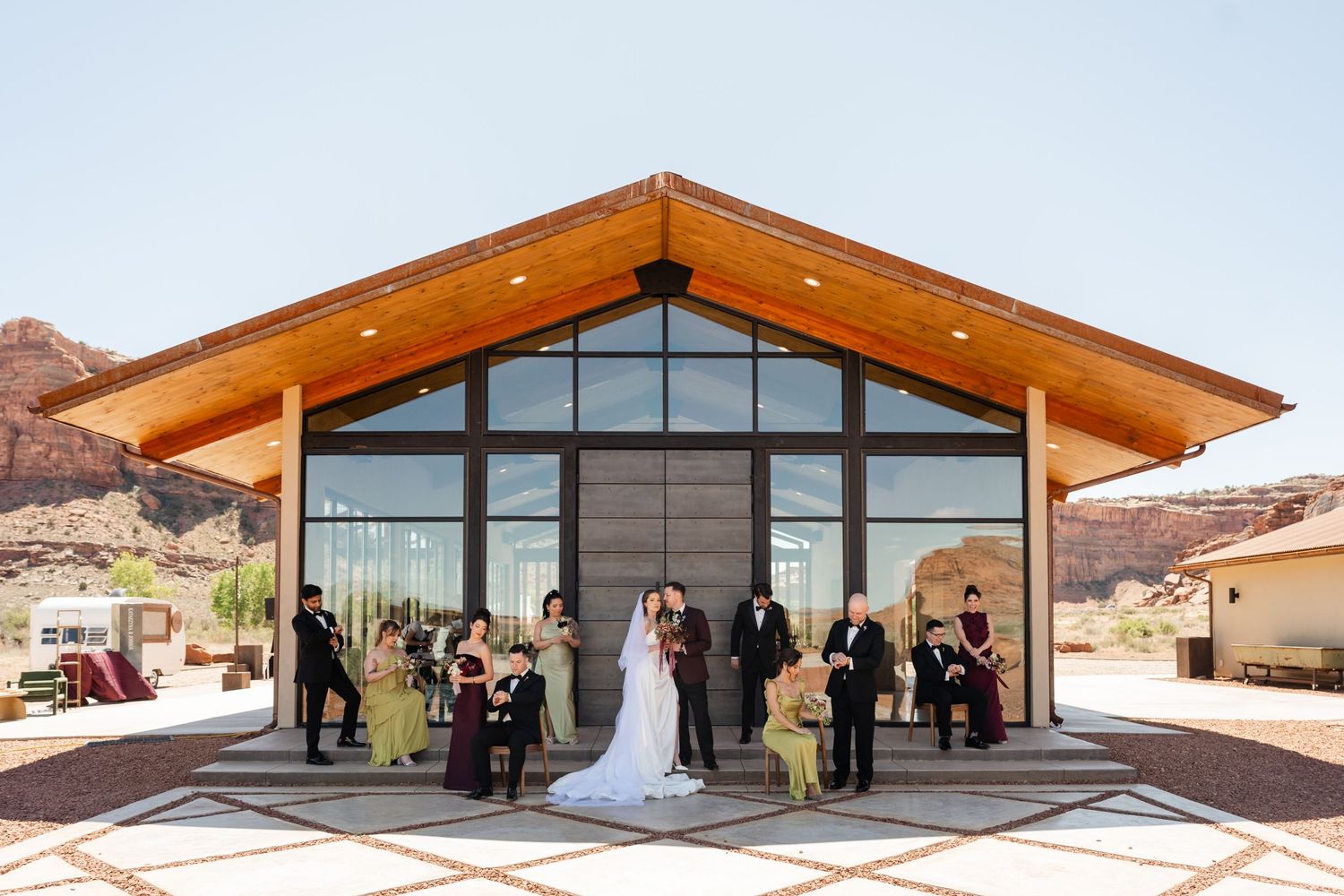 Wedding ceremony outside modern desert venue with red rock mountains, bridal party in burgundy and gold dresses, sunny blue sky.