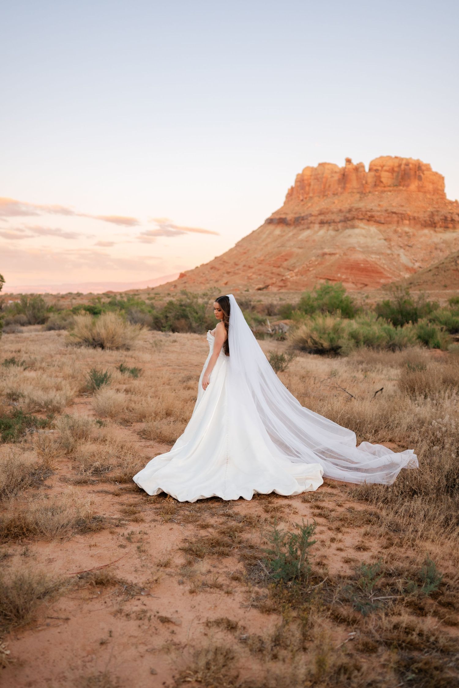 Bride in white strapless gown with long cathedral veil flowing behind her stands before glowing red rock butte at sunset.