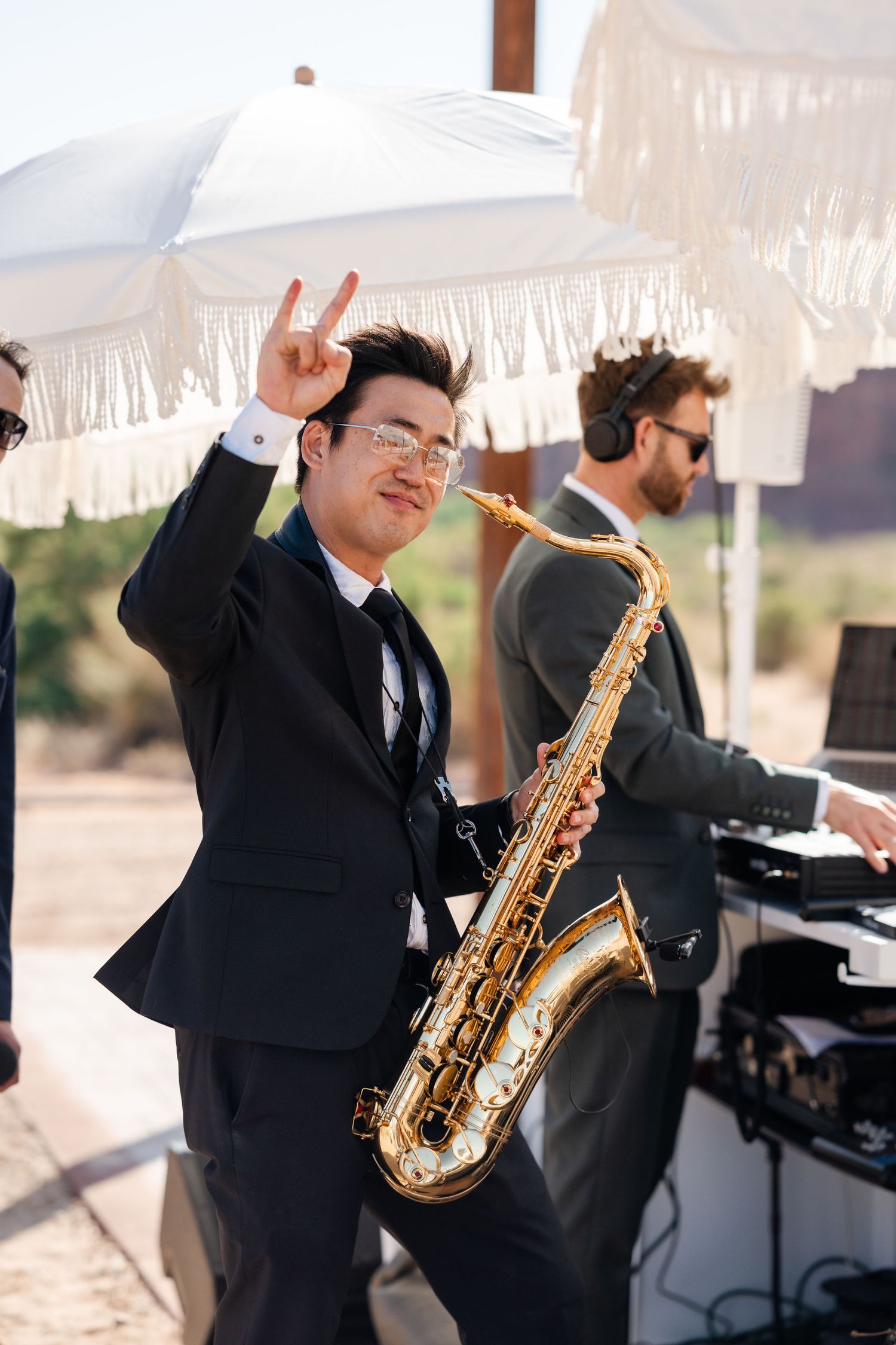 Saxophonist in black suit making rock sign at outdoor wedding reception with DJ setup and white fringed umbrella.